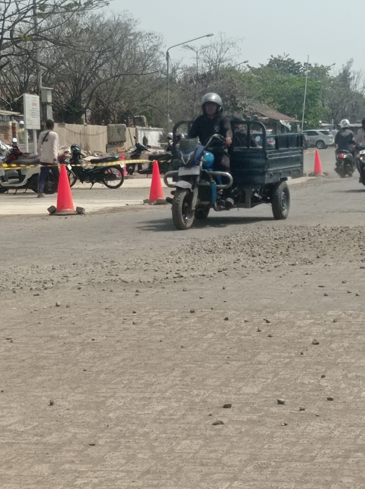 An eye-level, medium shot captures a street scene in Tegal, Indonesia, with a focus on construction or roadwork. A three-wheeled vehicle, likely a motorcycle with a cargo bed, is central, driven by a person wearing a helmet. The vehicle is dark green and blue, with a metal cargo bed. The driver's face is partially obscured by the helmet. In the background, a white car is visible, along with other motorcycles, one of which is being ridden by a person in a helmet. Several orange traffic cones are placed on the ground, suggesting a demarcated area or hazard. The road surface is rough, with loose gravel and dirt, indicating ongoing work. Trees with bare branches are seen in the background, along with a building. The overall atmosphere suggests a busy, active work zone.