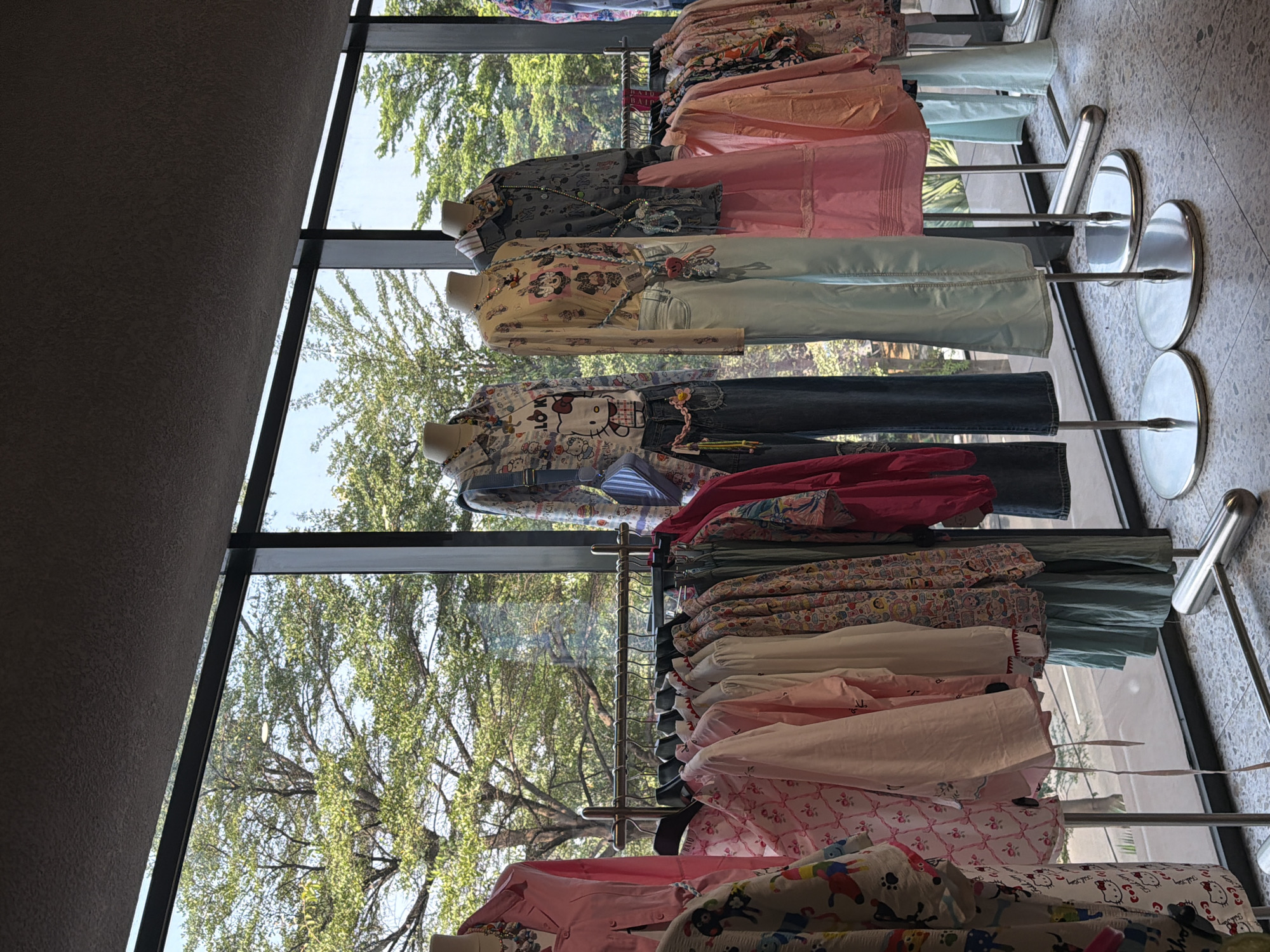 The image depicts a retail display of women's clothing inside a store. Several mannequins are dressed in colorful outfits, including shirts with cartoon prints and wide-leg pants in pastel colors. Garments are also hung on racks in the foreground, featuring similar prints and vibrant hues. The store appears to have large windows overlooking a lush, green outdoor environment with trees and sunlight. The flooring is tiled, and metal clothing racks and display stands are visible. The scene is static and there are no people actively engaged in any actions within the frame. The location is identified as East Jakarta, Indonesia.