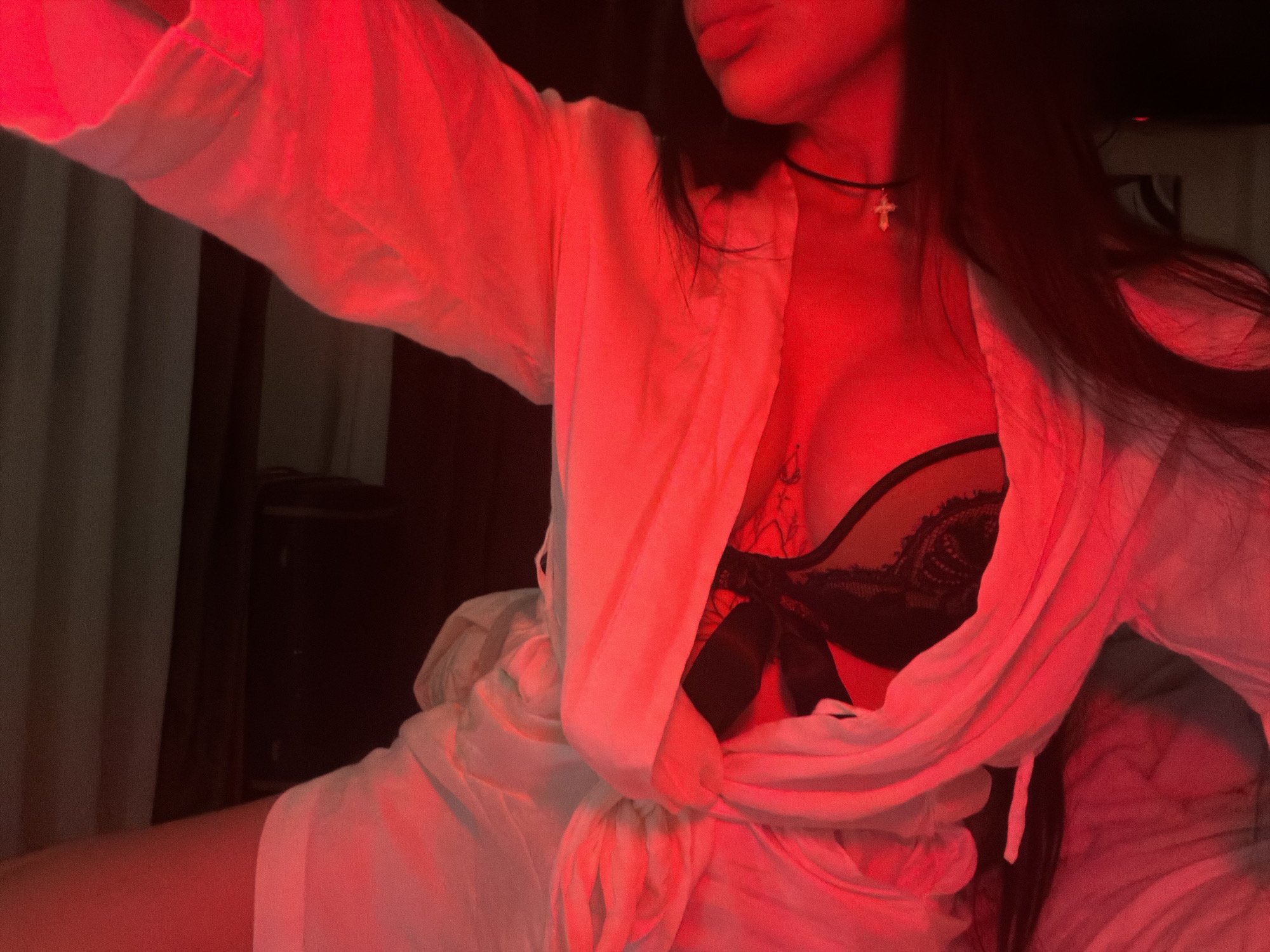 A woman with long dark hair and light skin is partially visible in a red-toned photograph. She is wearing a black lace bra, a white button-down shirt tied at the waist, and a black choker with a cross pendant. Her upper body is angled towards the left, with her left arm extended. Part of her left leg is visible. A small, dark tattoo is visible on her upper chest. The background is dark and indistinct, showing glimpses of furniture and curtains. The location of the image is not discernible as Boreti, Montenegro.