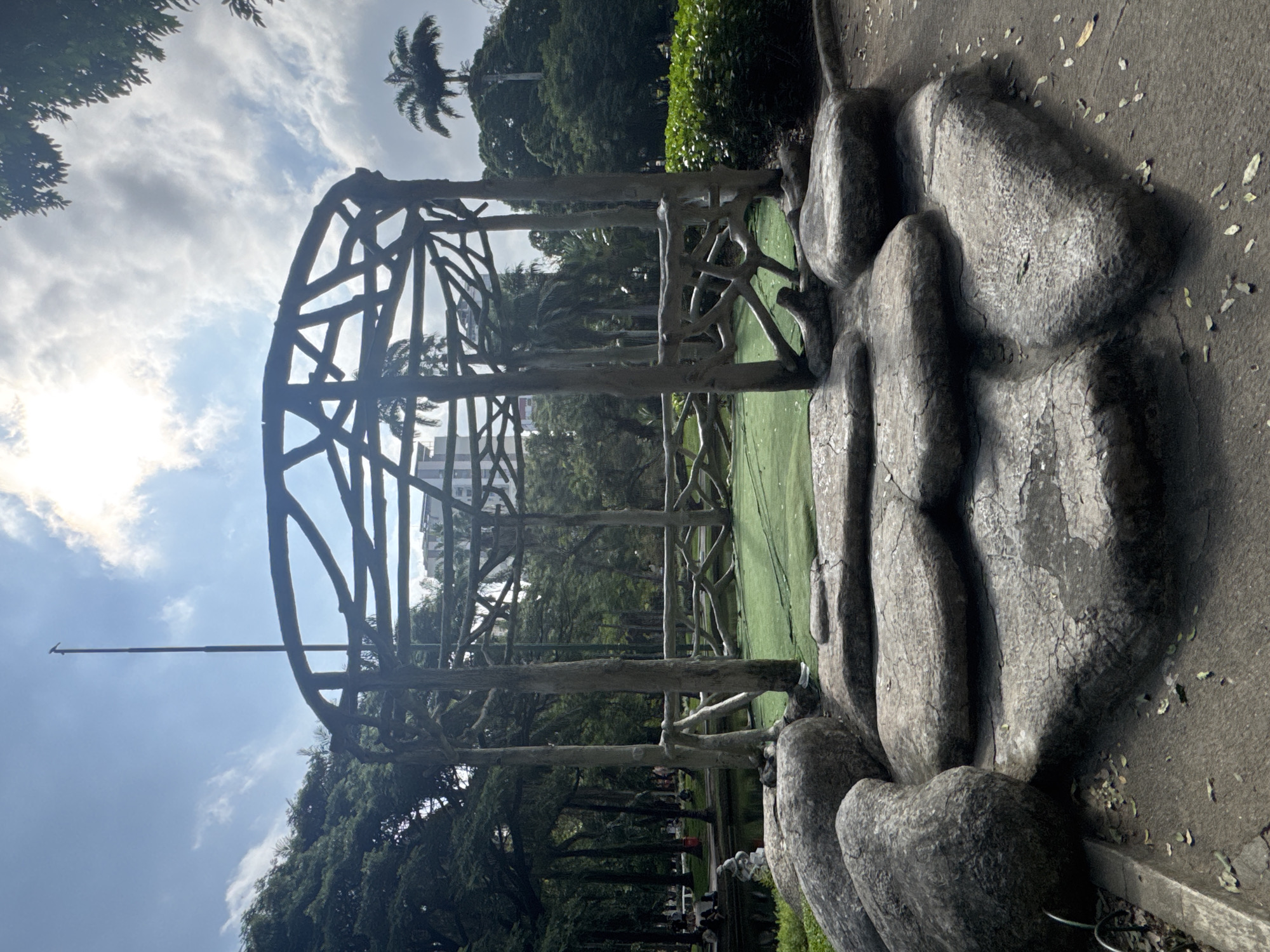 The image depicts a park scene in Niterói, Brazil, on a partly cloudy day, likely in the morning or early afternoon, as indicated by the bright sunlight and scattered clouds. The primary subject is an abstract, skeletal wooden structure that resembles a gazebo or a large birdcage, constructed from interwoven branches and logs. This structure is situated in the foreground, to the left of the frame, and is partially obscured by a series of large, rounded, grey, stone-like forms. These forms, possibly decorative elements or part of the park's landscaping, are textured and appear weathered, with some cracks visible.
Behind the wooden structure, a patch of vibrant green artificial turf is visible, suggesting a recreational area or a visually striking garden feature. Beyond the turf, a dense arrangement of trees forms a lush backdrop, with palm trees interspersed among them, a common sight in tropical and subtropical regions like Brazil. In the distance, a modern building with multiple stories can be seen peeking through the trees, providing a subtle contrast between the natural elements and urban development.
A tall, thin pole, possibly a flag pole, extends vertically into the sky from the left side of the image, its tip slightly angled. The overall atmosphere is tranquil and suggests a leisurely day in a public park. There are some indications of people in the background, further back in the park, mostly blurred and indistinct, engaged in passive activities like sitting or walking, contributing to the sense of a peaceful public space. No visible text is present in the image.