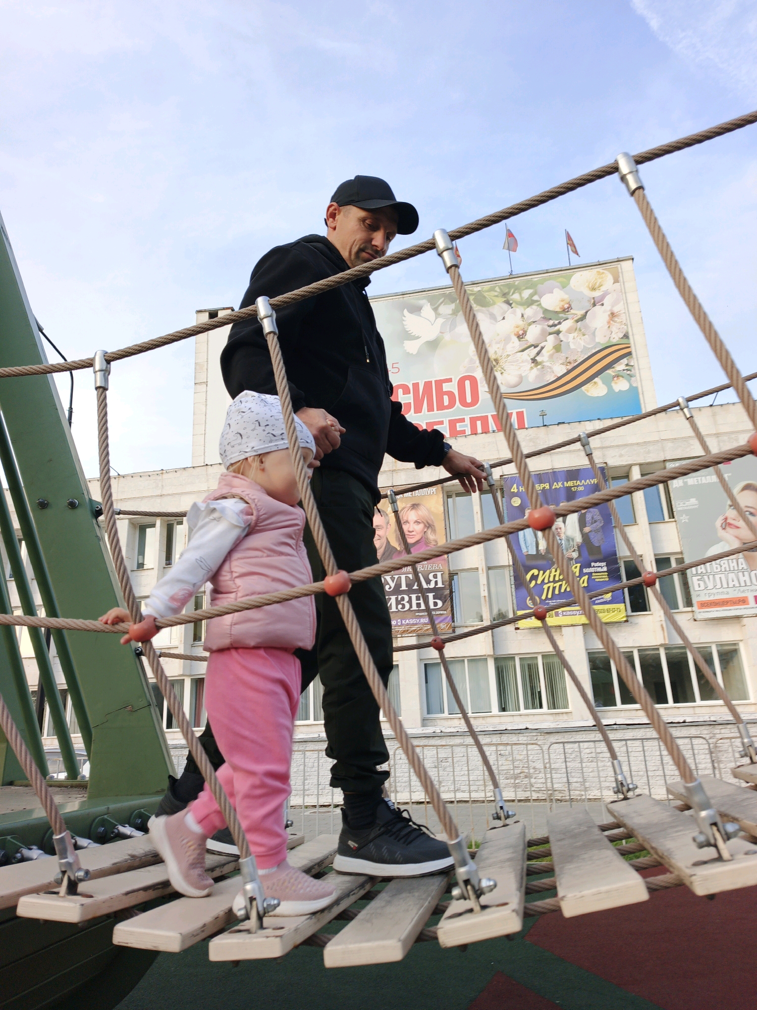 In Zlatoust, Russia, a man wearing a black baseball cap and a black hoodie, along with black pants and black sneakers, is walking on a rope bridge with a young child. The child, wearing a white bandana with black penguin imprints, a pink vest, and pink pants, is also on the rope bridge. The man is holding the child's hand, and both are looking forward. Behind them, a building with several posters is visible. One large poster features flowers and a dove, with Russian text. Other posters display people and text, some of which appear to be advertisements or event announcements. The sky is partly cloudy. The rope bridge has wooden planks for steps, connected by thick ropes.