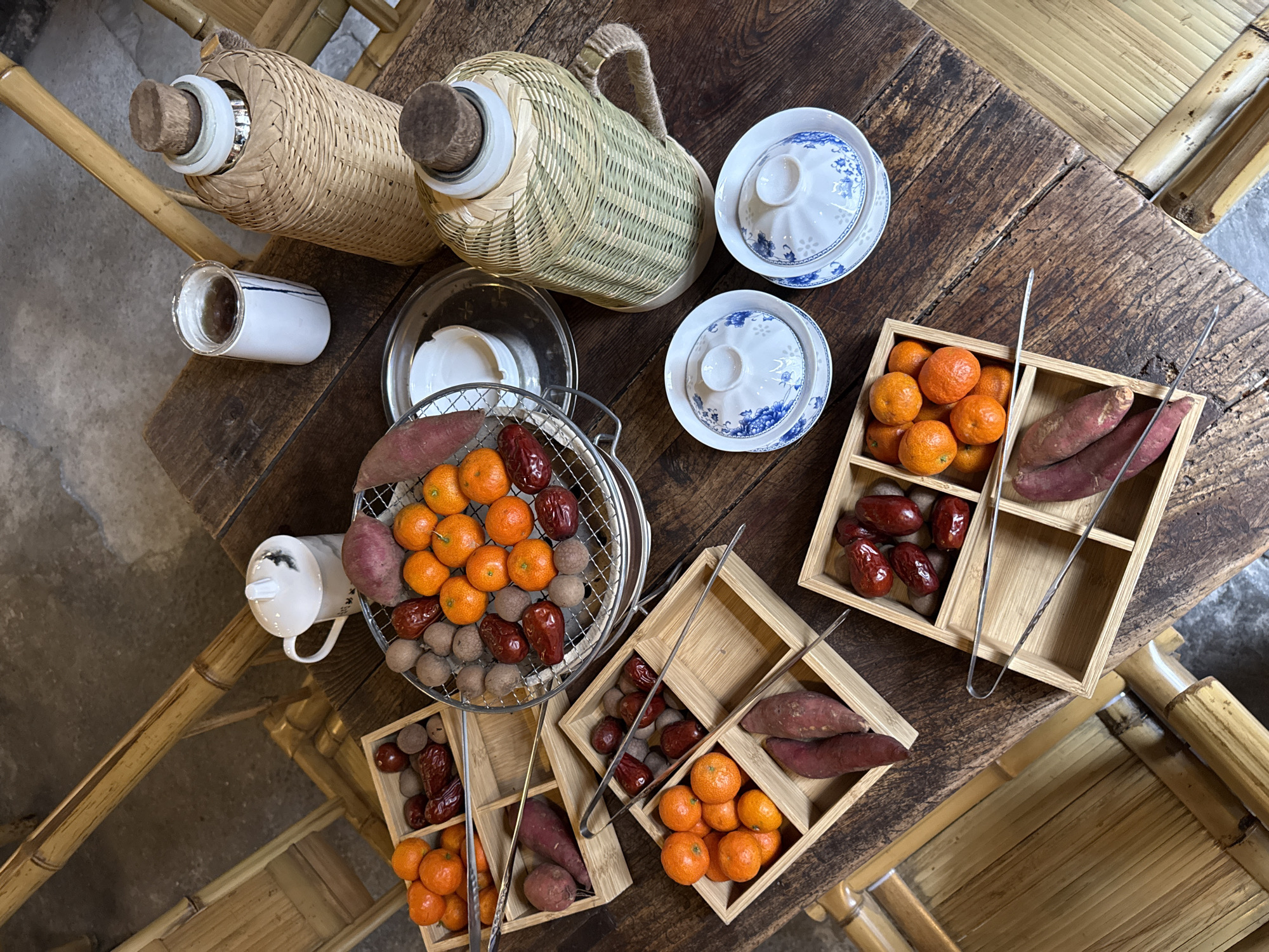 The image displays an overhead view of a rustic wooden table set with various food items and beverages, situated in Yuzhong District, China. The dark brown wooden table exhibits visible grain and plank construction. Sections of natural bamboo furniture, likely chairs or benches, are visible around the table's perimeter.
Central to the table is a round metal grill containing an assortment of small orange mandarins, dark red dates, purple-skinned sweet potatoes, and small, light brown, round fruits resembling longans.
Arranged on the table are four square, light-colored bamboo trays, each divided into compartments. These trays also hold combinations of mandarins, red dates, sweet potatoes, and longans. Each tray is equipped with a pair of metal tongs or skewers.
Beverage service includes two tall, insulated bottles, each encased in woven bamboo or rattan and sealed with a cork stopper. One bottle features a lighter, natural bamboo weave, while the other displays a darker, green-toned weave. Additionally, two white ceramic lidded cups are present; one has a dark liquid visible. Two traditional white porcelain teacups with blue floral patterns and matching lids (Gaiwan style) are also on the table. A small white ceramic dish rests on a silver-colored tray.
The floor visible in the background is concrete. No individuals are present in the scene.