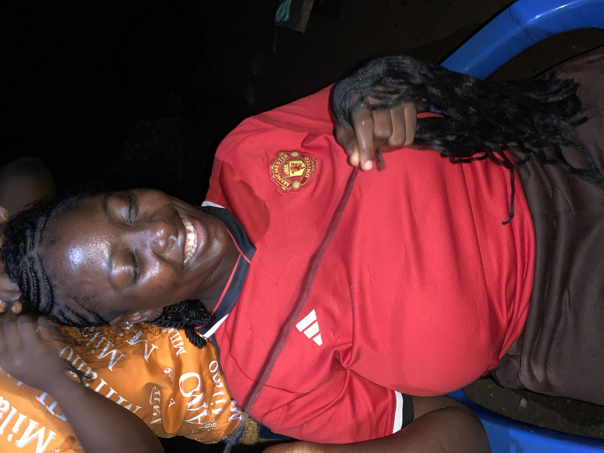 In Akwanga, Nigeria, a woman with dark skin is seated outdoors in a blue plastic chair, undergoing a hairstyling process. She is wearing a red Manchester United jersey with black and white trim and a brown skirt. Her eyes are closed, and she is smiling, with sweat visible on her forehead and cheeks. Her hair is being braided, and a portion of the braided hair is held in her right hand. Another person's hands are visible above her head, actively braiding her hair. The background is dark, suggesting the photo was taken at night or in a dimly lit area. Part of an orange t-shirt with the word "Milano" printed on it is visible behind her. A plastic cutting tool and some green scissors are on a wooden surface in the background.