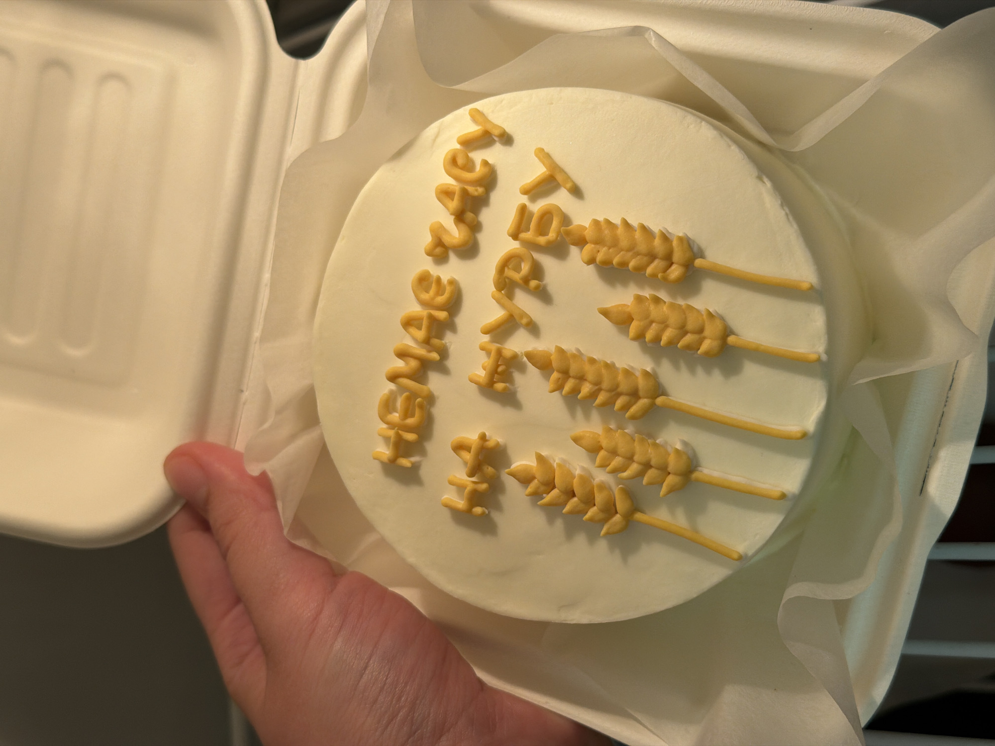 A person is holding a small, round, white cake in a white styrofoam takeout container. The cake is decorated with yellow icing letters that read "Have a great day" arranged vertically. Three wheat stalks, also made of yellow icing, are positioned beside the words. The cake appears to be resting on a sheet of white tissue paper inside the container. The location in the city of Tel-Aviv, Israel is not discernible from the image.