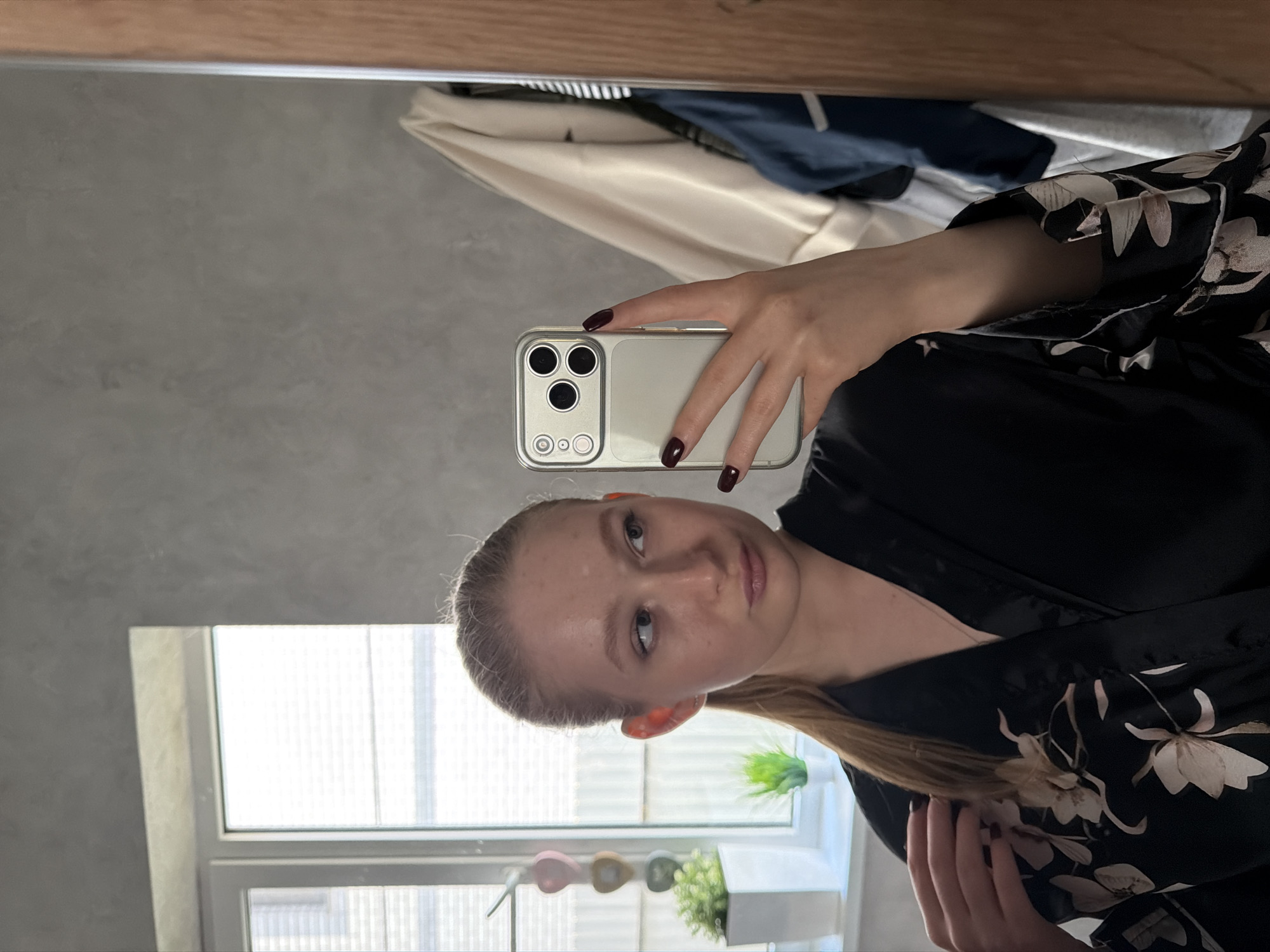 A light-skinned person holds a smartphone up in front of themselves, presumably taking a selfie. The smartphone's back is visible, revealing three cameras. The person has light blonde hair pulled back. The person is wearing a dark robe with a floral pattern. In the background, there is a window with a plant in a pot on the sill. There is also a mirror with clothing hanging behind the person. The background wall is grey. The location is not discernible.