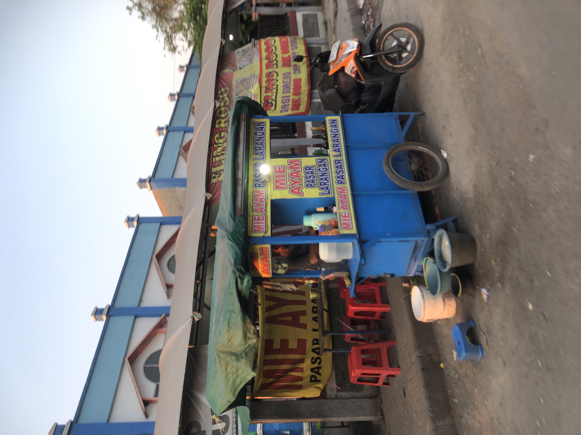 The image shows a street scene in Larangan, Indonesia, with a focus on a blue food cart. The cart is labeled "MIE AYAM" and "PASAR LARANGAN", indicating it sells chicken noodles in the Larangan market. A green tarp covers the top of the cart. Red plastic stools are arranged in front of the cart. To the right of the cart, a black and orange motorcycle is parked. In the background, a building with a blue and white facade is visible, along with banners advertising "ELING ROSO". Several buckets are placed on the ground near the cart. The sky is clear and appears to be dusk.