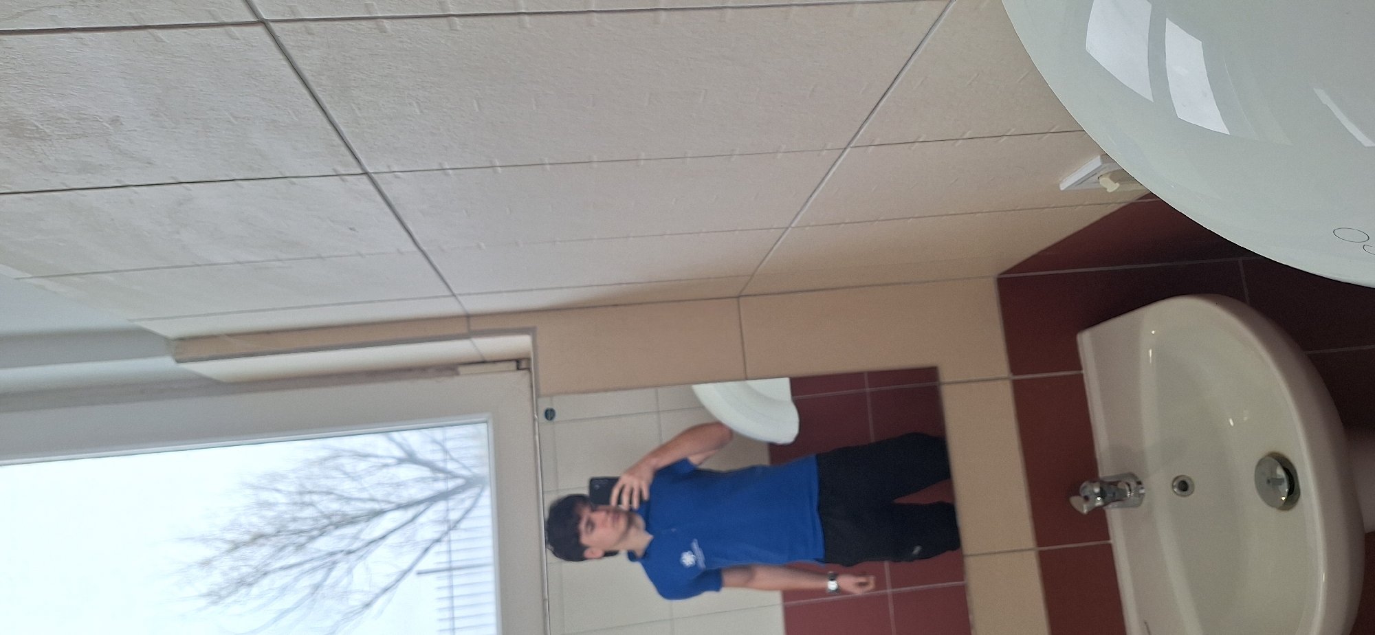 A photo taken inside a public restroom, likely located in Burgas, Bulgaria, is oriented at an angle, showing a tile floor and ceiling. A window is on the left side of the frame revealing a bare tree against a cloudy sky. The reflection of a young man taking the photo is visible in a mirror. The man is wearing a blue shirt and black pants. A white sink and toilet are on the right side of the frame, set against brown and burgundy tiled walls.