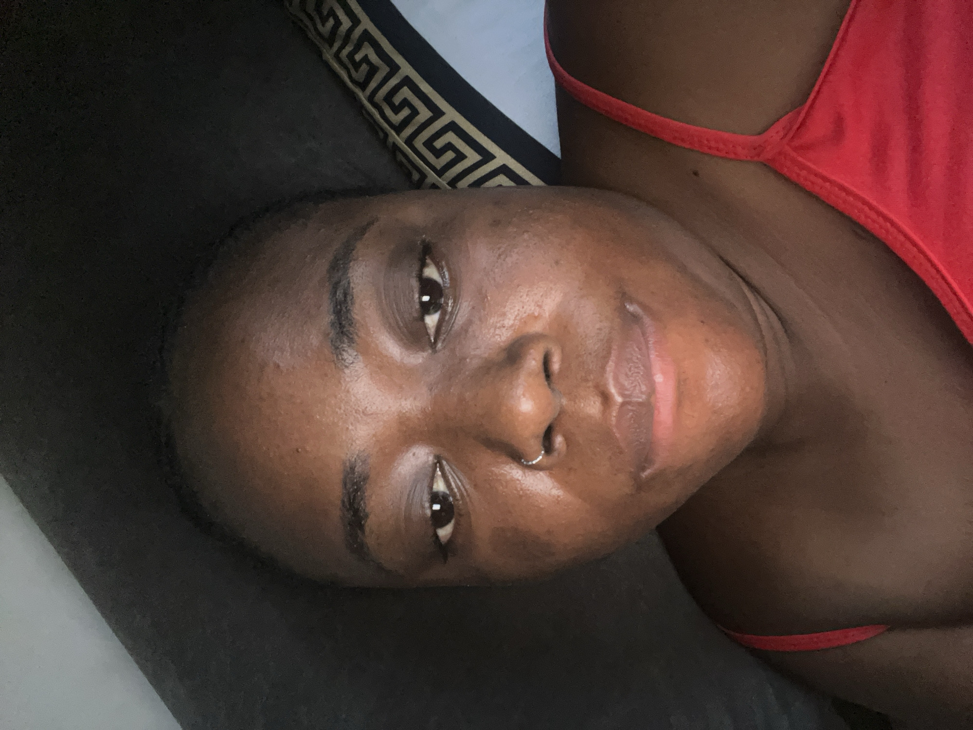 A close-up portrait shows a young adult woman lying down, looking directly towards the camera with a soft, relaxed expression and a subtle smile. She has a dark skin tone, full lips, and dark eyes with visible reflections, indicating an indoor light source. A silver nose ring is present on her right nostril. She is wearing a red top, with a thin strap visible over her right shoulder.
The background consists primarily of dark-colored fabric, likely bedding, suggesting an indoor setting such as a bedroom. A patterned pillow is visible in the upper right quadrant, featuring a black and gold geometric design resembling a Greek key pattern. The overall mood of the image is calm and comfortable. The provided context indicates the location is Abuja, Nigeria, though no specific visual cues within the image confirm this. There is no legible text visible in the frame.