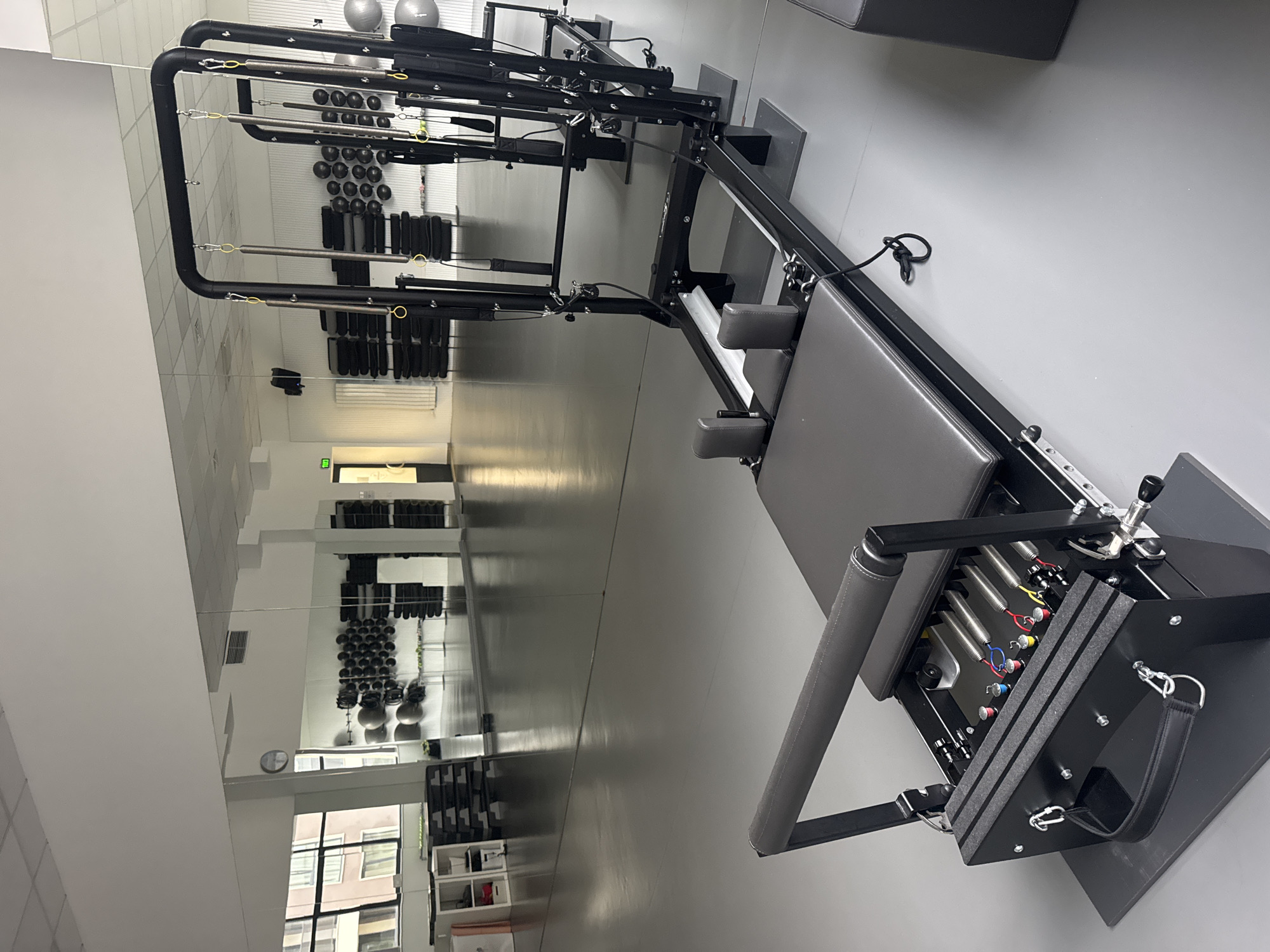The image shows a Pilates reformer machine in a mirrored fitness studio. The reformer is black with a gray carriage and headrest. Springs of various colors are visible at the base of the machine. The studio has a gray floor and white walls. In the reflection in the mirror, racks of dumbbells and other fitness equipment are visible, along with a window showing buildings outside. The location is not identifiable as Sofia, Bulgaria from the visual information provided.