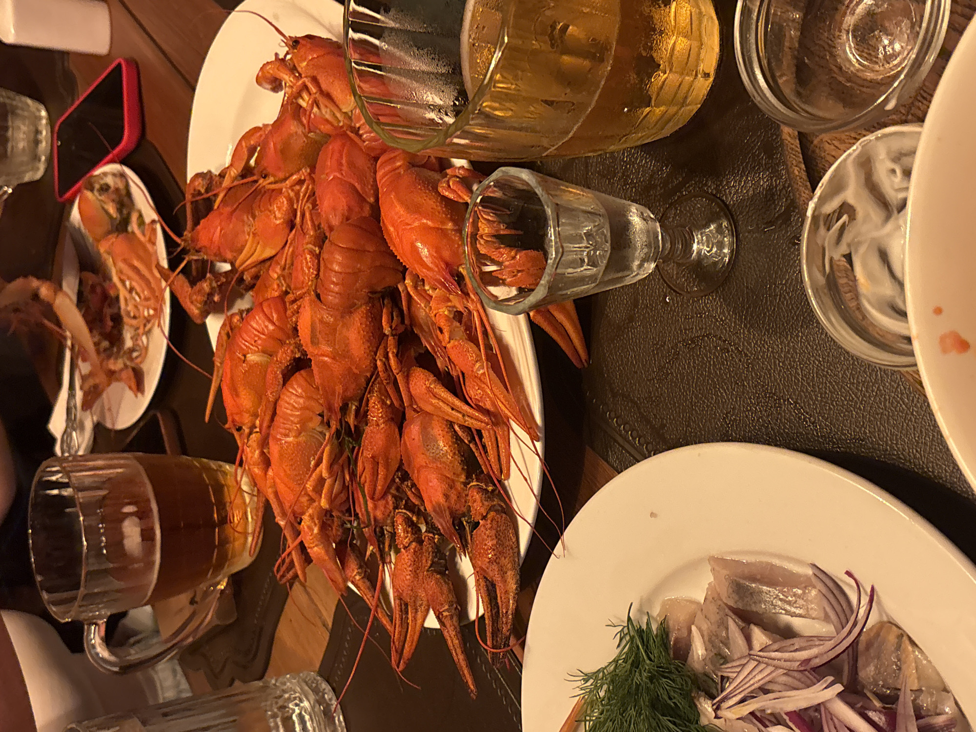 A table laden with food and drinks. A large white plate dominates the scene, piled high with cooked crawfish, dyed red. Next to it is a plate of herring with sliced red onions and dill sprigs. Various glasses and mugs hold beverages, seemingly beer and vodka. A small plate on the side contains more crawfish. A red-cased smartphone rests next to that plate. The surface beneath appears to be leather, suggesting a higher-end establishment. The location is likely Krasnoyarsk, Russia given the cuisine.