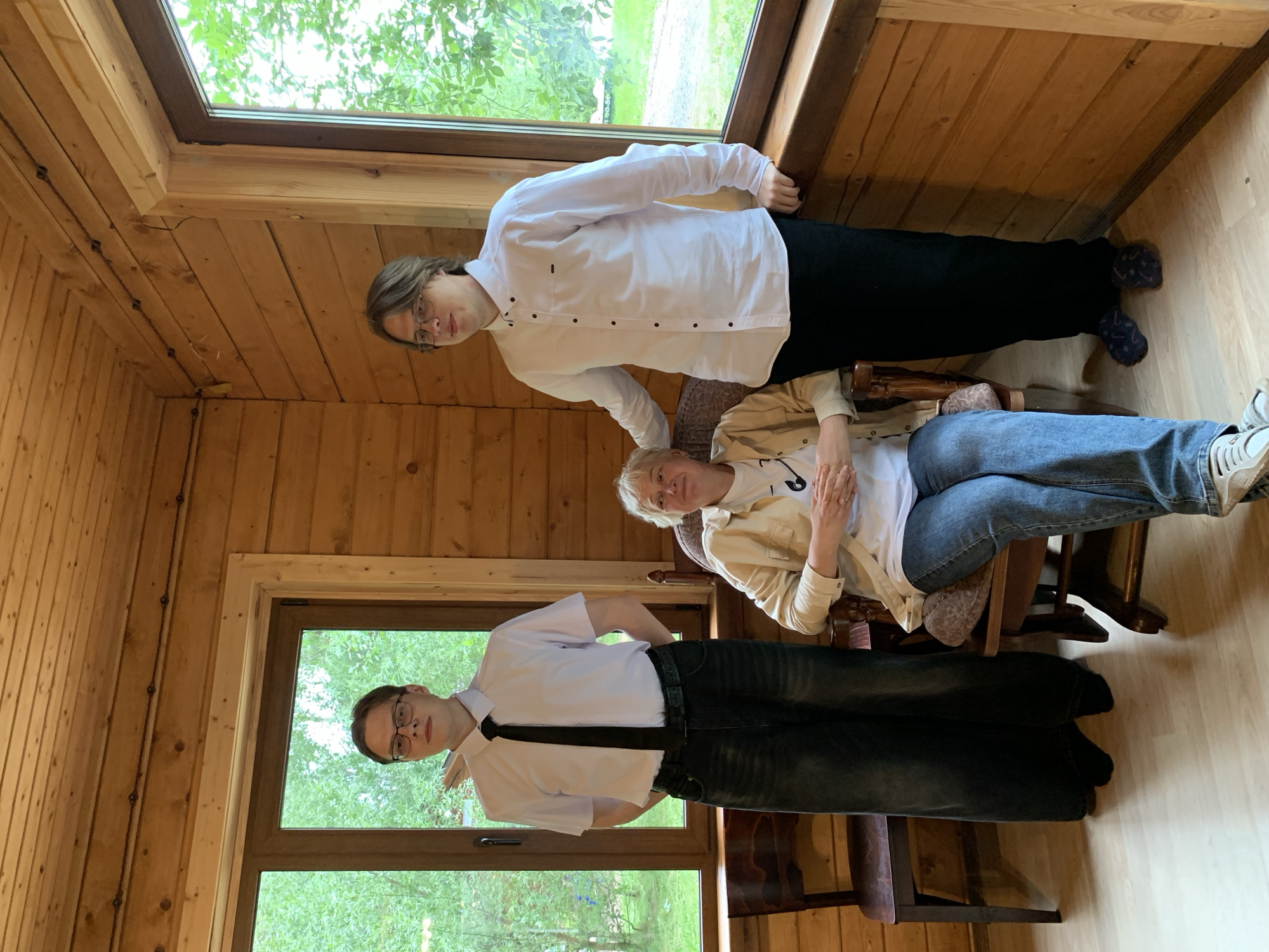 The image depicts an indoor scene within a room constructed of wooden planks. The room features large windows overlooking a green, tree-filled exterior. In the foreground, a woman is seated in a wooden rocking chair, wearing a light-colored jacket over a white t-shirt and blue jeans. Two young men stand in the background. The man on the left is wearing a white short-sleeved shirt with a black tie and dark, wide-legged pants. The man on the right is dressed in a white long-sleeved shirt and black pants. The overall ambiance suggests a casual gathering in a rustic setting. The location is identified as Chaikovsky, Russia.