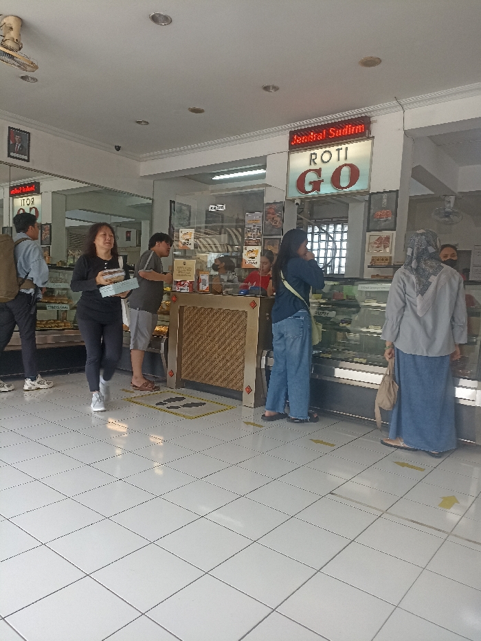 The image shows the interior of a bakery called "Jendral Sudirman Roti Go" located at the coordinates -7.428, 109.247 in the city of Purwokerto, Indonesia. The bakery has a tiled floor with yellow tape arrows directing customers. There are five people inside: two customers waiting at the counter, one customer near the counter to the left, one cashier, and one person standing further back to the left. There is a large display case with prepared goods in the center of the image, while the counter is on the left. The bakery has a white ceiling with recessed lighting and a sign with the name of the bakery in red lettering. The walls are white and there are several posters on the wall behind the counter. There is a large window to the right of the display case showing a glimpse of the street outside. A person in the back is wearing a mask. There are some bags on the counter near the display case. The bakery is well-lit.