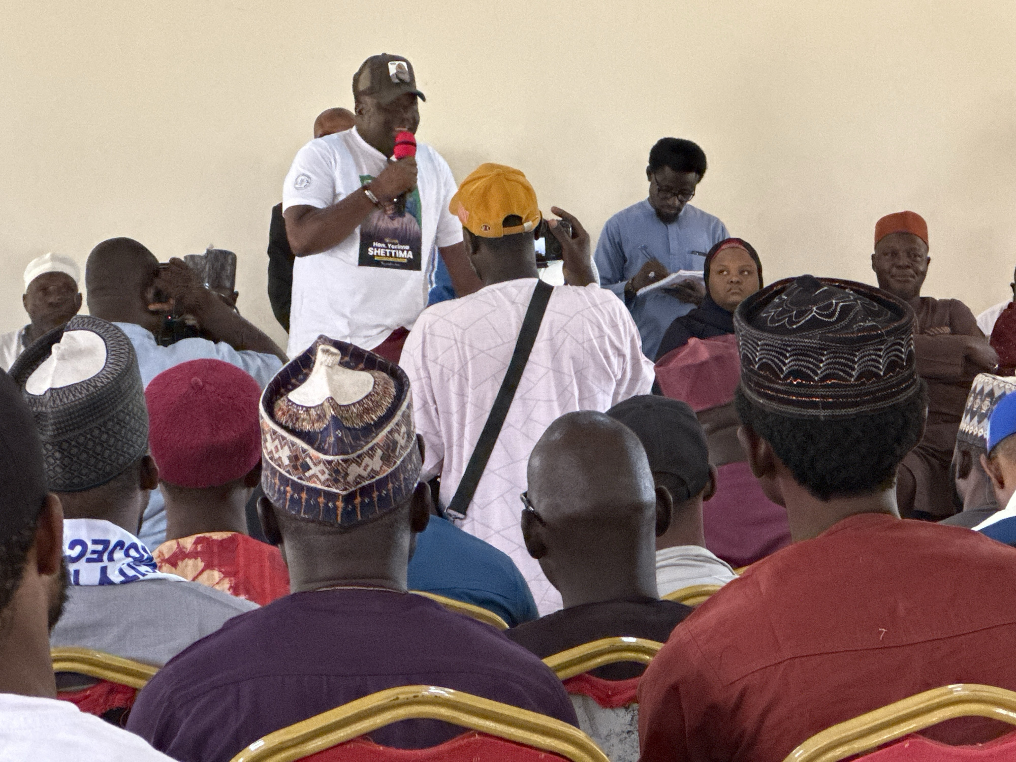 In Unknown, Unknown, a speaker stands before an audience. He holds a microphone and wears a white t-shirt, and a baseball cap. A person in a yellow cap is in the process of taking a photo. A man in glasses takes notes, and a woman in a headscarf is seated in the audience. The audience is diverse in its attire, with many wearing traditional hats and clothing. The environment appears to be an indoor meeting space.