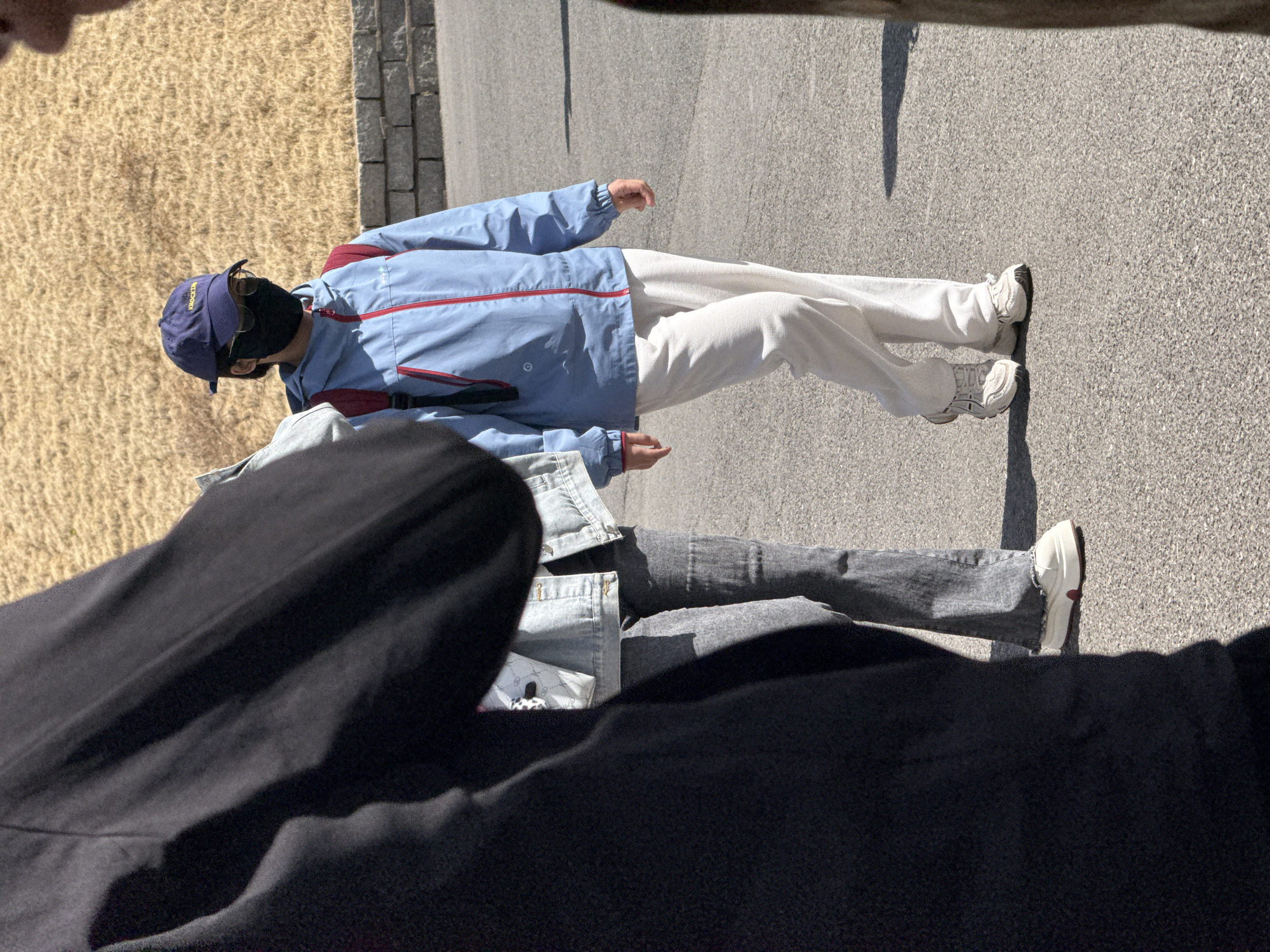 A photograph taken in Zhongtun, China shows two figures walking on a paved surface next to a grassy area. The first figure, who is wearing a navy cap, sunglasses, a black face mask, a light blue jacket with red trim, and white pants, is fully visible. The second figure, who is wearing a denim jacket and gray jeans, is partially obscured by a large black object in the foreground. The lighting suggests it is daytime.