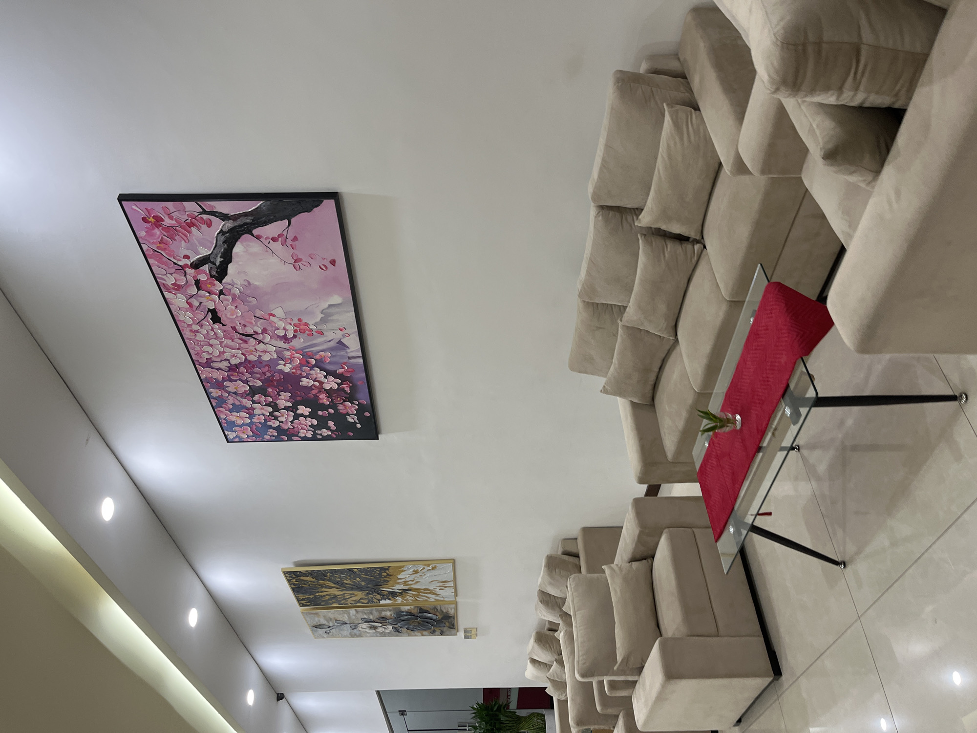 In the city of Binong, Indonesia, a waiting area features a light-colored sectional sofa with several throw pillows. In front of the sofa is a glass coffee table with a red runner and a small potted plant. Two pieces of abstract art hang on the white wall above the sofa. The floor is tiled with large, light-colored tiles. The lighting appears to be recessed ceiling lights and an indirect cove light.