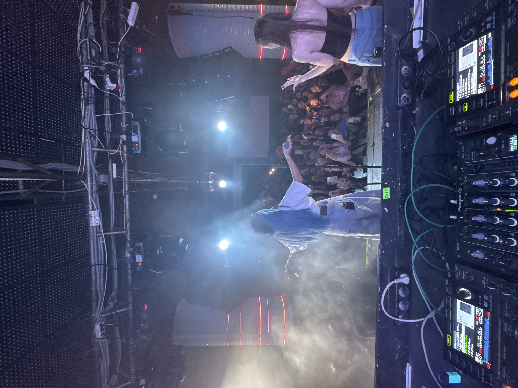 The image shows a DJ performance at a concert. The location is likely in Kazan, Russia. There are two DJ's and a large crowd. The DJ on the left has blue hair, white shirt, and light jeans. The DJ on the right has long hair and a black top and jeans. Visible equipment includes a DJ board, lighting equipment, a disco ball, and a sound system. The crowd is dense, with many people holding up phones. The stage is illuminated by bright spotlights. There is a decorative lighting structure in the background, emitting red light. Cables and wires are visible along the left side of the image.
