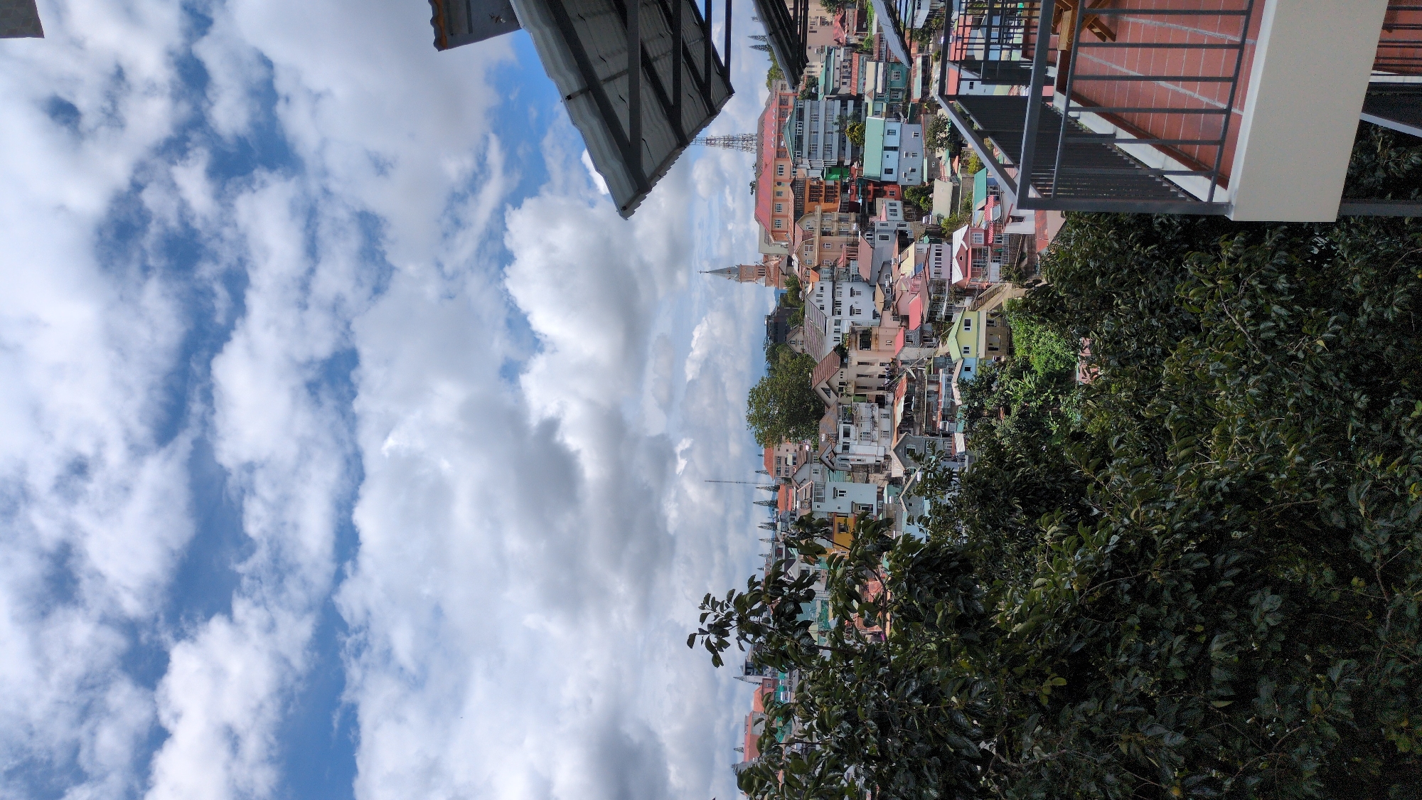 This is a vertical, eye-level, medium shot of a densely packed cityscape in Da Lat, Vietnam, viewed from a balcony. In the foreground, dark green foliage from trees obscures the left side of the frame. To the right, a balcony with terracotta tile flooring and a dark metal railing is visible. Beyond the trees, the city unfolds with numerous buildings of varying heights and colors, including houses and structures with pitched roofs. Notable structures in the midground include a prominent bell tower of a church and a tall antenna mast. The sky above is filled with scattered white clouds against a blue background. The overall impression is of a vibrant, hilly urban landscape under a partly cloudy sky.