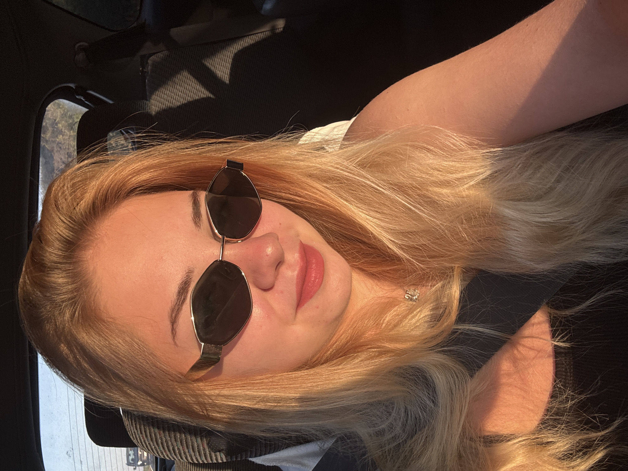 A young woman with long, blonde hair and sunglasses is sitting in the passenger seat of a car. The car appears to be in motion, as evidenced by the blurred background and the sunlight casting shadows. The woman is looking towards the camera, and her lips are slightly parted, suggesting she might be speaking or smiling. She is wearing a black tank top and a silver necklace. The interior of the car is visible, with the headrest and seat of the driver's seat in the background. The overall lighting suggests it is either early morning or late afternoon, with the golden hour sun illuminating the scene. The context of Sredets, Bulgaria, is not visually apparent in the image itself; the scene depicts a common car interior setting.