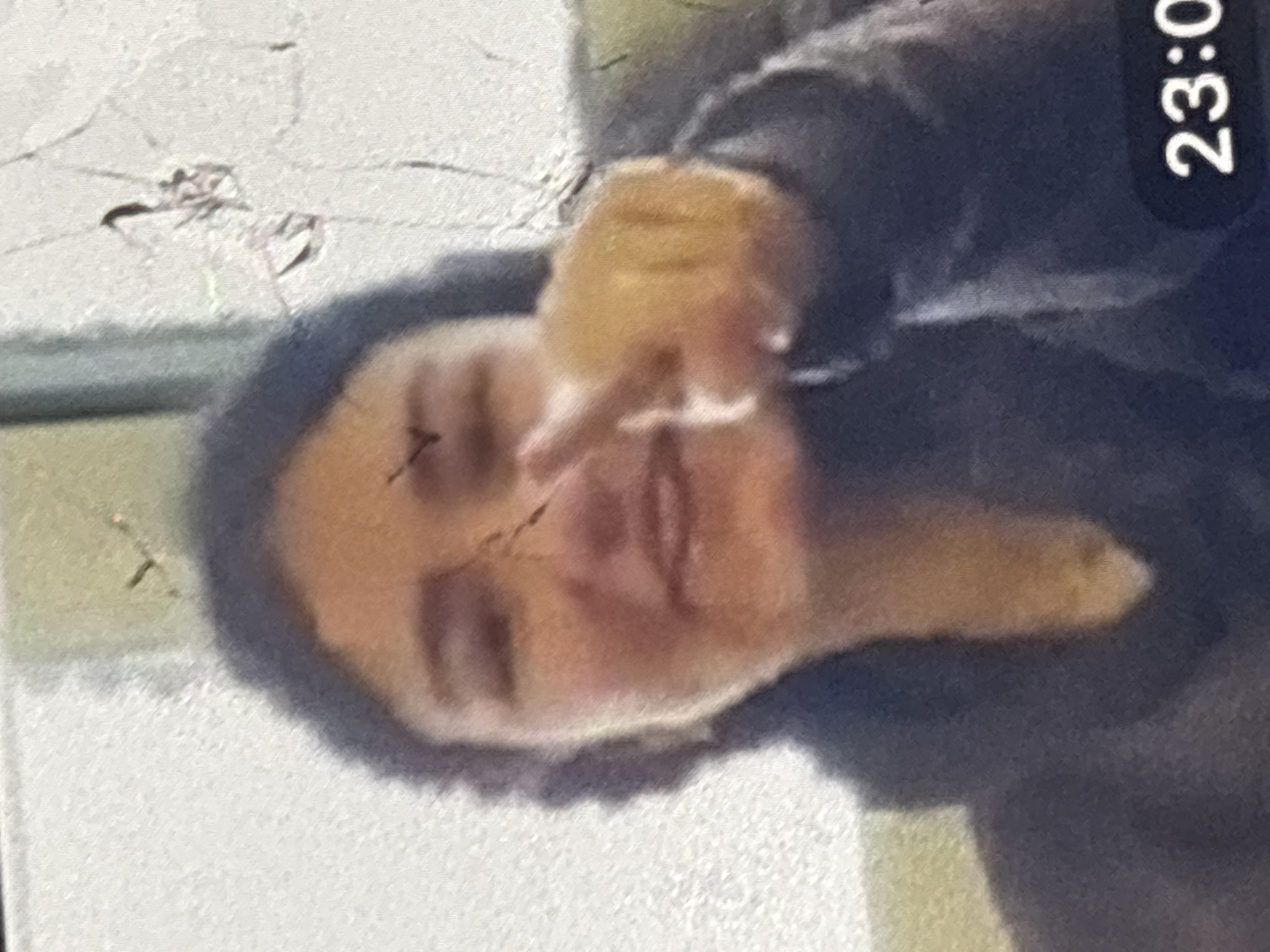 The image is a low-resolution, pixelated close-up of a young person's face, looking towards the camera. The person has dark hair and is wearing a dark hoodie. Their right hand is raised, with a finger pointing directly at the camera. The background is blurry and appears to be an indoor setting, possibly a room. There is a timestamp in the bottom right corner displaying "23:0". The image quality is poor, with visible scratches and artifacts on the screen, suggesting it may have been taken from a device screen. There is no visible information in the image that would allow for an identification of the specific location within the city of Orihuela, Spain.