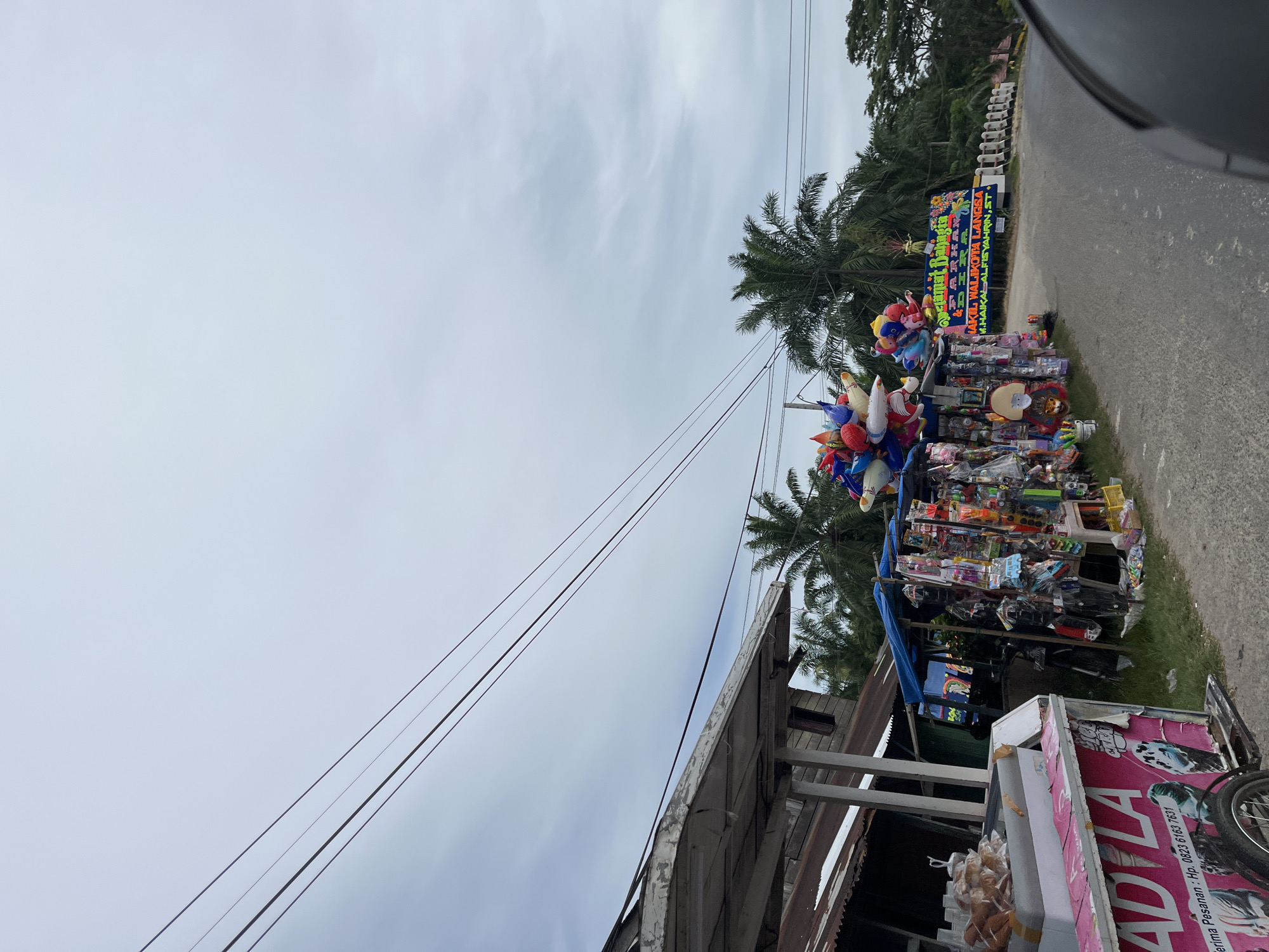 The image depicts a roadside scene in Seunebobaru, Indonesia, with a focus on two street vendors. To the left, an ice cream cart, predominantly pink and white, displays the text "ADILA" and a phone number, and contains what appears to be bread rolls in a cooler. Adjacent to the ice cream cart is a stall selling various small items, including balloons, toys, and other merchandise, displayed on racks and a table. Further down the road, another stall or display is visible with a blue tarp overhead, showcasing more goods. In the background, palm trees line the road, and a sign with Indonesian text is prominently displayed. Power lines stretch across the overcast sky. The road is paved, and the environment appears to be a rural or semi-rural area. No people are clearly visible in the image.