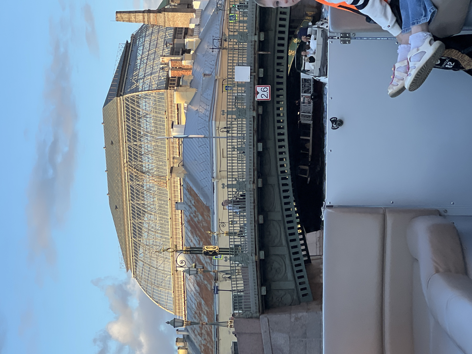 The image shows a view from a boat in Saint Petersburg, Russia, looking towards a large, arched glass-roofed building and a bridge. The sky is blue with scattered clouds. The foreground shows the interior of the boat with seating and a person's legs. The bridge is green with decorative elements and has a sign with "2.6" on it. Several other boats are visible on the water beneath the bridge, carrying passengers. The large building in the background appears to be a public structure, possibly a market or exhibition hall, with a distinctive glass roof. The overall scene captures a moment of a boat tour on a sunny day in Saint Petersburg.