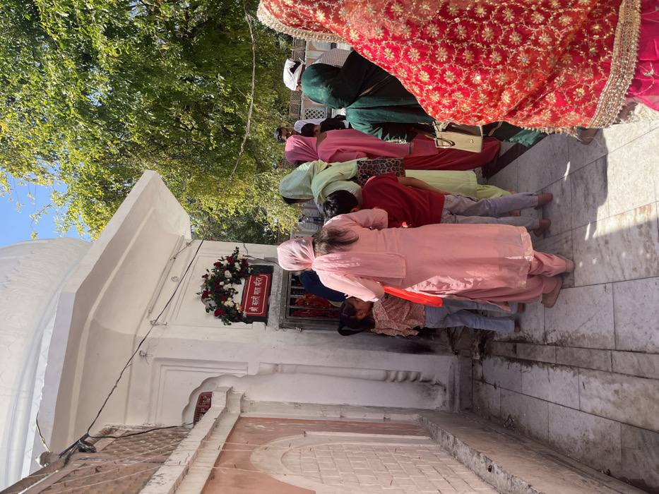 The image shows a group of people standing in front of a small white building with a dome. The building appears to be a temple, as evidenced by the floral garland above a sign that reads "Shri Kali Maa Mandir". The building is located in Panchkula, India at the coordinates 30.723 latitude and 76.86 longitude. There are several women and children in the group. They are mostly wearing traditional Indian clothing. The women are all wearing headscarves, and some of them are wearing long, colorful dresses. The children are wearing a variety of clothing. The people are standing on a white tiled walkway, and there are some plants and trees visible in the background. The image appears to depict a scene of religious devotion.  A man in a white shirt and blue jeans is standing on the left side of the building.  A woman in a pink dress is in the center.  A woman wearing a red and gold sari is on the right side of the photo.  A person in a green garment is standing in front of the woman in the red sari.  There is a red bag visible on the ground.
