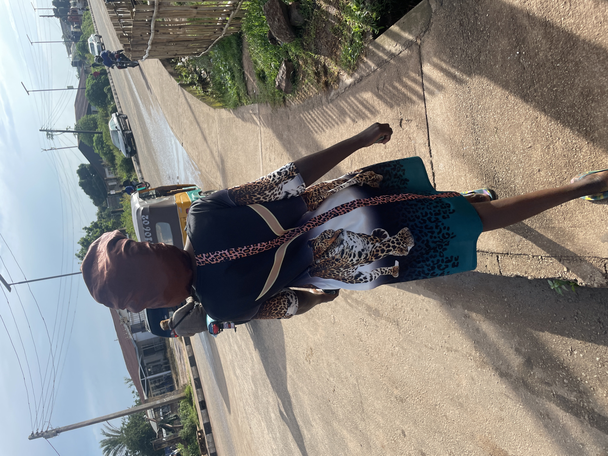 A young person wearing a satin bonnet and a leopard-print dress walks on a sidewalk beside a road in Jos, Nigeria. The person is walking away from the camera. Behind them, a yellow auto-rickshaw, a motorcycle with a rider, and a few cars are visible on the road. The environment appears to be a city street with buildings and vegetation in the background. The sun is casting long shadows on the sidewalk.