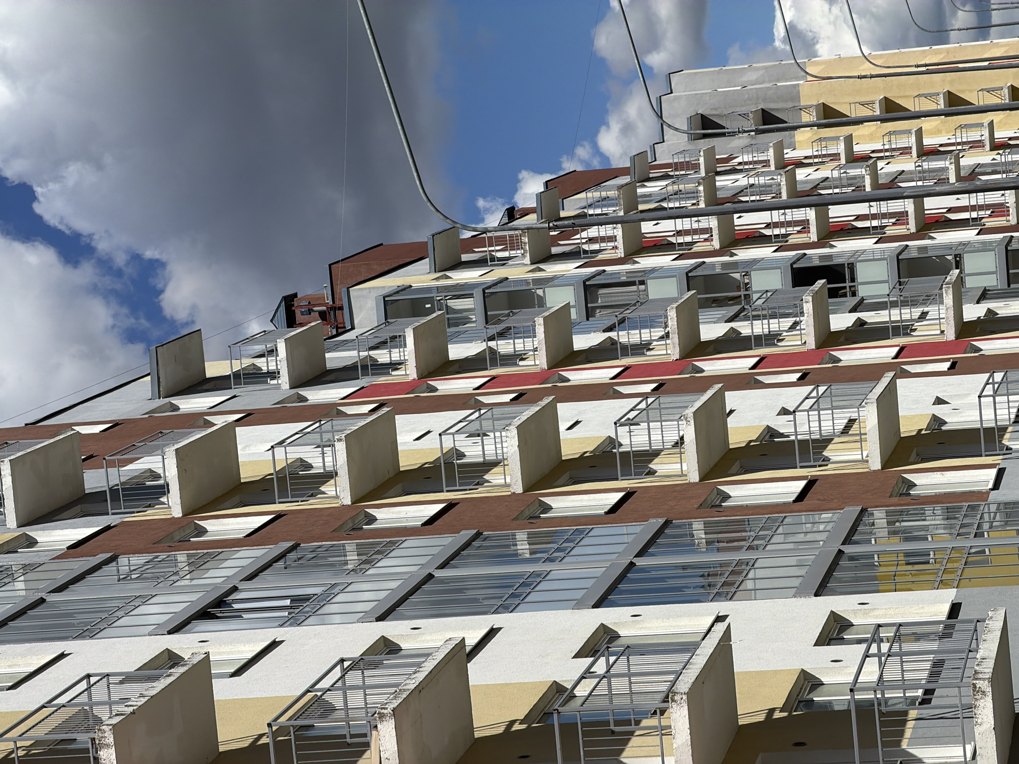 The image shows the facade of a multi-story building in Янино-2, Russia. The building has a stepped design, with each level slightly recessed compared to the one below. Each level features a row of identical rectangular balconies with minimalist metal railings and concrete dividers. The building's exterior is painted in horizontal stripes of white, yellow, brown, and red. Small windows are also visible between the balconies. The sky is partly cloudy, with blue patches visible behind the building. A metal cable runs across the upper part of the image.