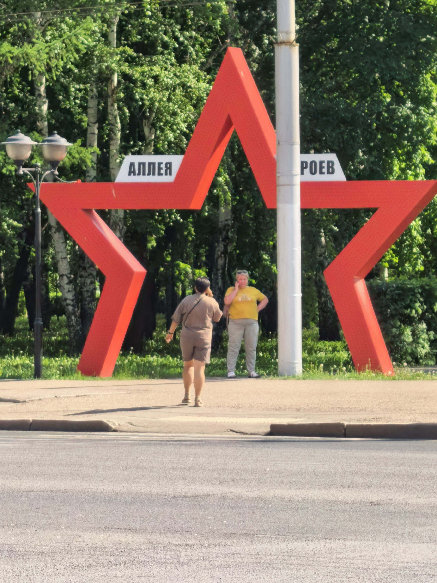 The image captures an urban scene in Sterlitamak, Russia. A large, bright red star structure dominates the foreground, with the words "АЛЛЕЯ" and "РОЕВ" affixed to it. This translates to "Alley of Heroes." A utility pole runs through the center of the star.
Two women are visible near the star. One woman, dressed in tan clothing and carrying a bag, is walking away from the camera and toward the star. The second woman, wearing a yellow shirt and light-colored pants, is standing near the star and holding a cell phone to her ear.
In the background, a dense array of green trees fills the frame, interspersed with occasional white birch trunks. A street lamp is visible on the left side of the image.
A street stretches across the bottom of the frame. A curb separates the street from the area with the star.