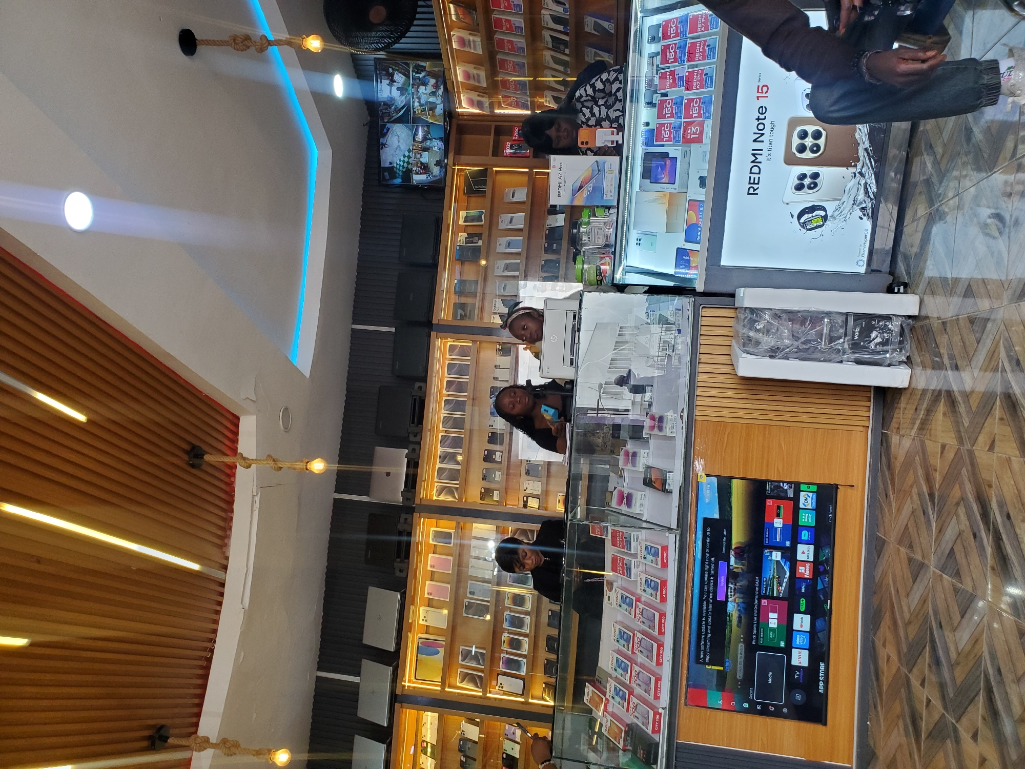 The image depicts the interior of a mobile phone and electronics store located in Jos, Nigeria. The store is well-lit with warm-toned lighting fixtures integrated into a wooden slatted ceiling and blue ambient lighting along a stepped ceiling feature. In the foreground, several display cases are filled with mobile phones and their packaging. On the left, a large television screen displays an interface with various app icons and a notification about a software update. Behind the display cases, three women are visible behind a counter, interacting with customers or staff. One woman in the center is wearing a black top and looking down. To her right, another woman with long braided hair is holding a blue mobile phone. On the far right, a fourth person, partially visible, is seated and appears to be a customer. The store shelves are stocked with numerous mobile phones and other electronic devices, arranged in an organized manner. The overall environment suggests a commercial retail space focused on consumer electronics.