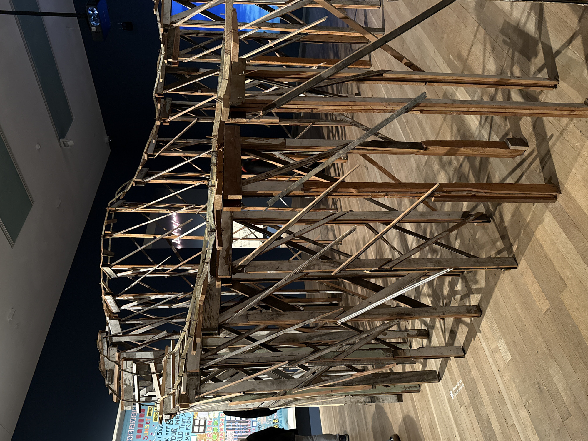 The image shows a complex wooden structure, appearing like a decaying architectural model or art installation, comprised of various wooden planks and beams arranged in an intricate, layered fashion. The structure casts shadows on a wooden floor. In the background, there's a wall with visible text and images, possibly part of an exhibition display. The setting appears to be indoors, potentially a gallery or museum space. The image does not provide enough information to identify a specific location in London.