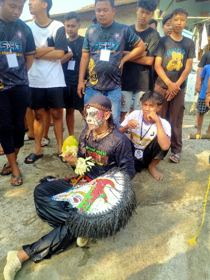 The image depicts a gathering of individuals in Sepat, Indonesia, outdoors on a paved or concrete surface under daylight conditions. In the foreground, a person is seated on the ground, wearing a black long-sleeved shirt, black pants, and light-colored shoes. This individual has elaborate facial makeup consisting of a white base with black outlining, red/pink accents around the eyes, and black web-like patterns on the cheeks and forehead. A dark headband is worn. They hold a light green, possibly young coconut, and several white flowers or leaves in their left hand. A large, ornate, fan-shaped or shield-like object with black fringe and a white surface bearing red, green, and black designs rests on the ground beside their right leg.
Around the seated individual, approximately eight to ten other males are present. Most are standing, while one young male crouches to the right of the seated figure, looking towards the camera. Many are wearing t-shirts, some with visible logos such as "PSHT" and "SIDOLUHUR" on lanyards or shirts, indicating potential affiliation with an organization. Attire includes various casual shirts, jeans, and shorts; footwear ranges from sandals to bare feet. The background shows parts of a simple building or wall and some sparse vegetation, consistent with a village or residential area. A yellow string or cord extends across the lower right of the frame.