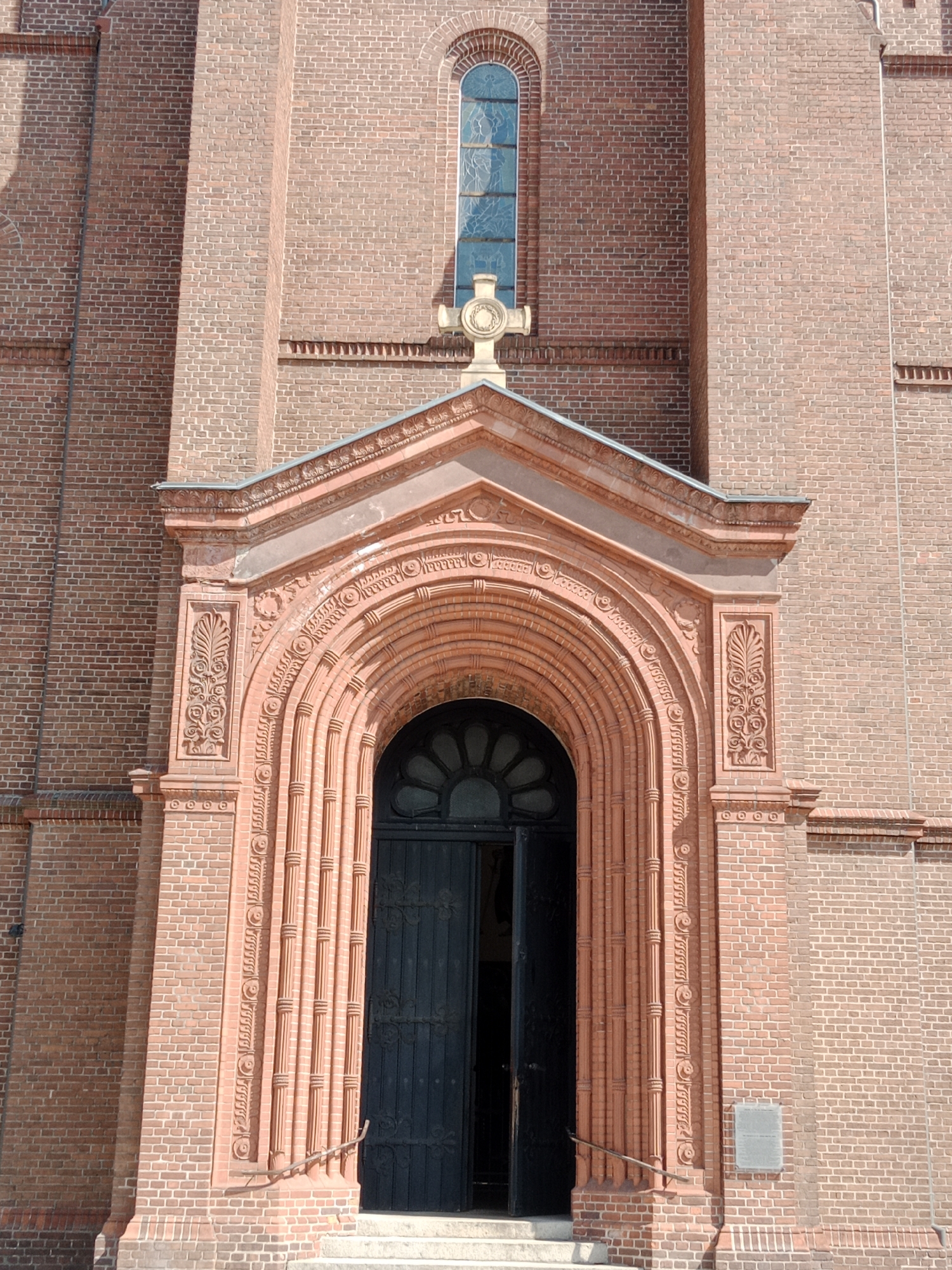 The image shows the entrance of a brick church. The entrance is an arched doorway with intricate carvings and a cross above. A tall, narrow stained-glass window is visible above the cross. The church is made of red brick, and the doorway is framed by ornate stone work. The doors are dark and open slightly, revealing the interior. The scene is brightly lit by sunlight, casting shadows on the building. The church is located in Bydgoszcz, Poland.