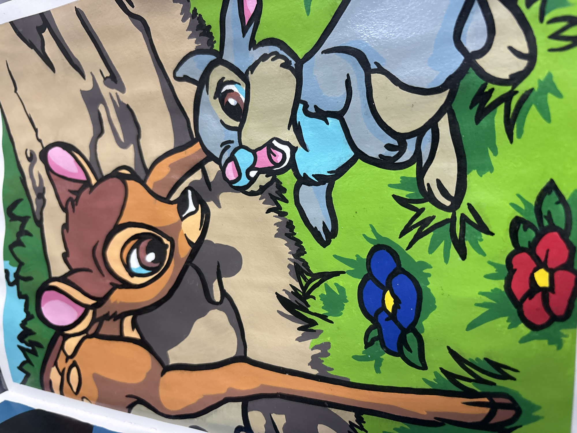 This is a close-up, stylized illustration of characters from Disney's Bambi. The main subjects are Bambi, a young deer, and Thumper, a rabbit. Bambi is depicted in the foreground, facing left with large, expressive eyes. His fur is a warm brown, and he has large, pink inner ears. He appears to be looking towards Thumper. Thumper is shown in the background, leaping or running. He is gray with blue accents and has wide, blue eyes and a pink nose. The characters are set against a backdrop of a tree trunk and green grass. There are also two stylized flowers in the grass: one blue with a yellow center, and one red with a yellow center. The overall style is simple and cartoonish, with bold black outlines and flat areas of color. The scene suggests a moment of play or interaction between the two friends in a forest setting. There is no visible text in the image. The location context of Omsk, Russia, does not appear to be directly indicated by any visual cues within the artwork itself.