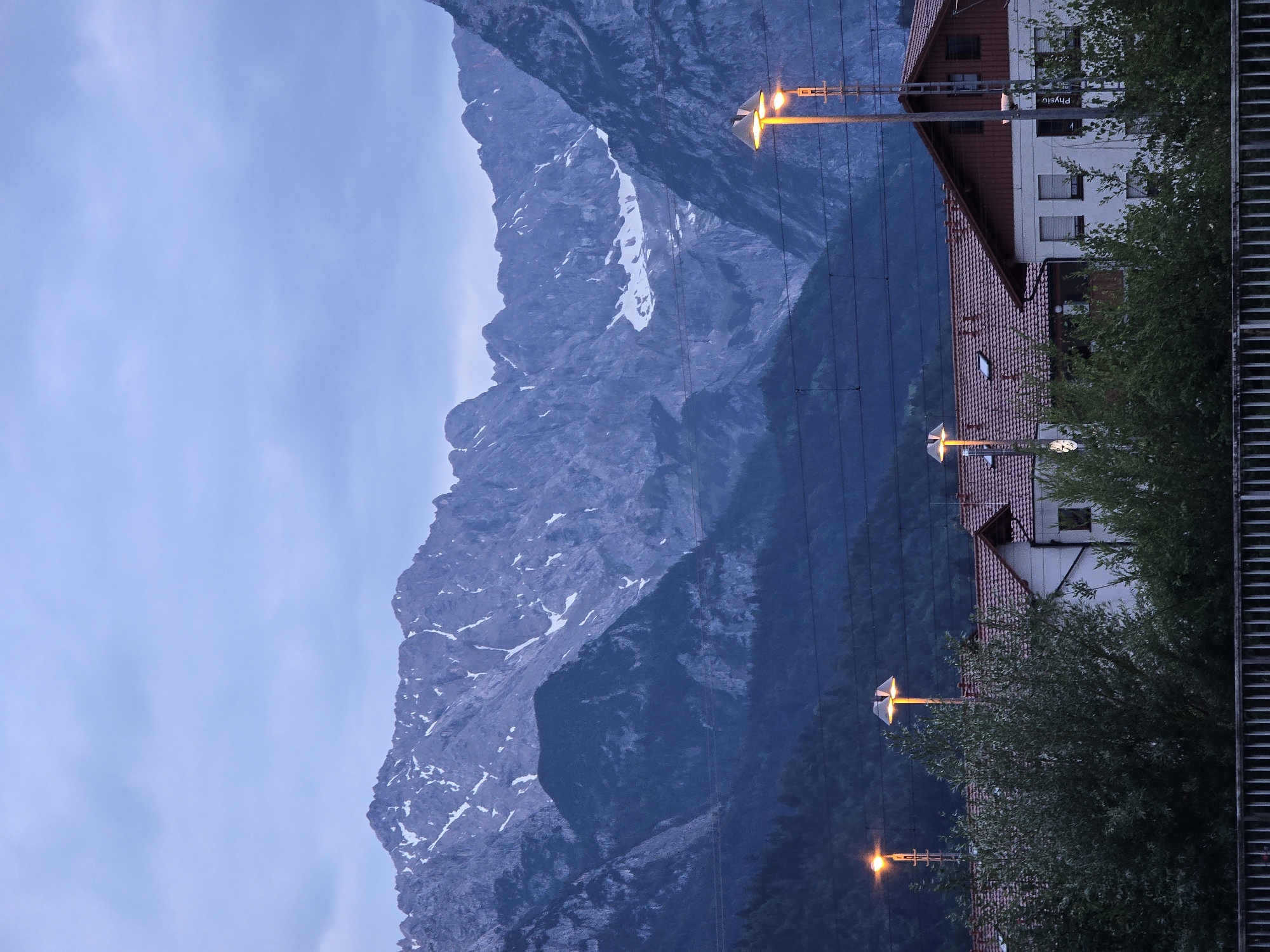 The image shows a view of the Zugspitze massif, the highest peak in Germany, as seen from Garmisch-Partenkirchen. The mountains are majestic and imposing, with snow-capped peaks and rocky slopes. In the foreground, there are a few buildings with red tile roofs, possibly part of the town or a nearby industrial area. There are also some trees and streetlights, suggesting an urban or suburban setting. The sky is overcast and the light is dim, indicating that it is either early morning or late evening. The overall impression is one of a dramatic and beautiful natural landscape, with a touch of human presence.