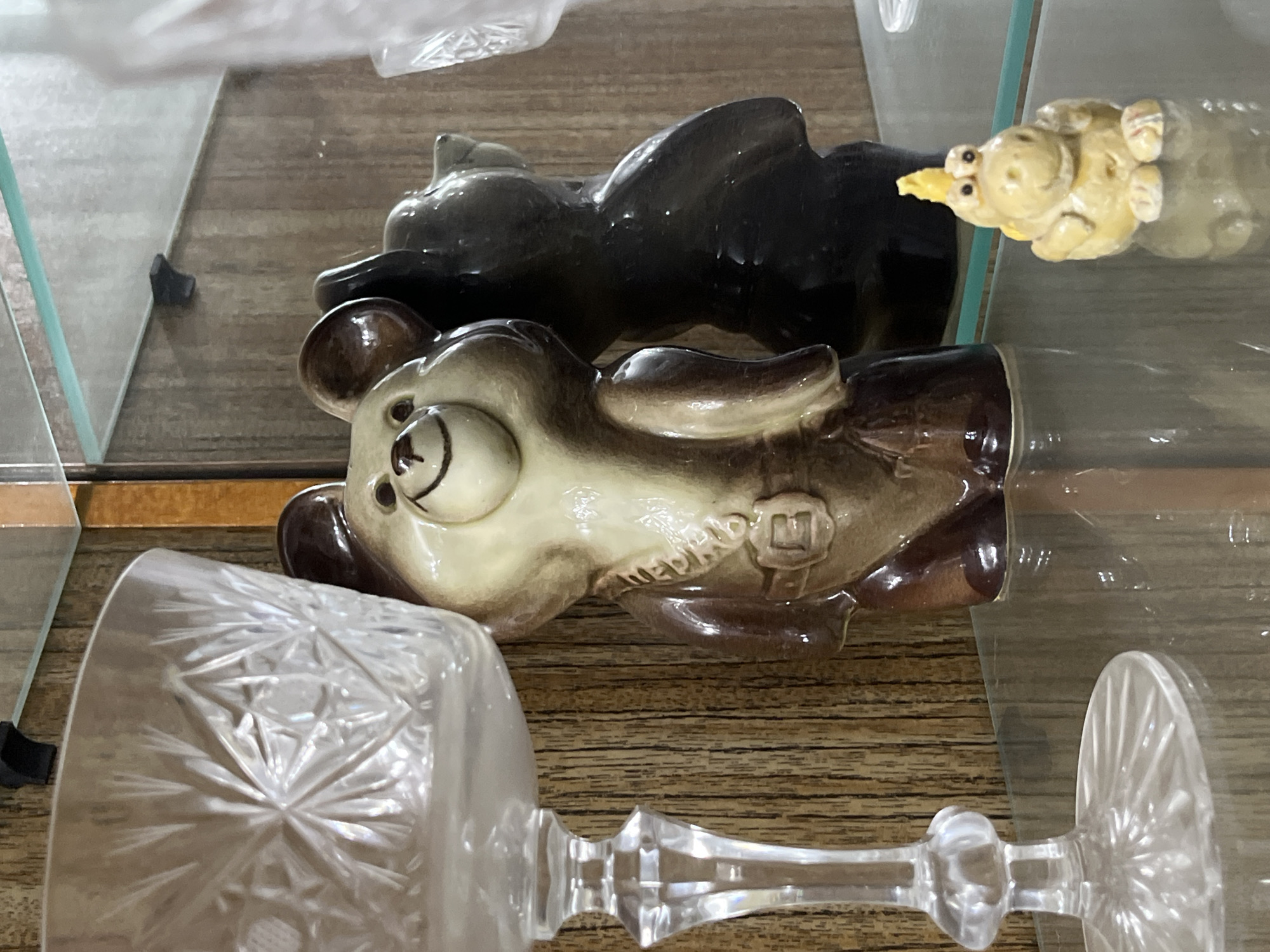A close-up, eye-level shot shows a display shelf containing various figurines and a cut-glass goblet. Two bear figurines are prominent: one in light brown and cream with visible Russian writing on its side, and another darker, almost black bear figurine lying behind it. To the right, a light yellow pig figurine is partially visible. In the foreground, the base and stem of a cut-glass goblet with a clear, detailed pattern are featured. The setting appears to be indoors, possibly within a display case in the city of Chaikovsky, Russia, given the Russian writing on the bear figurine. The shelf itself has a wood-grain surface. The photograph is well-lit, allowing for clear observation of the objects' textures and details.