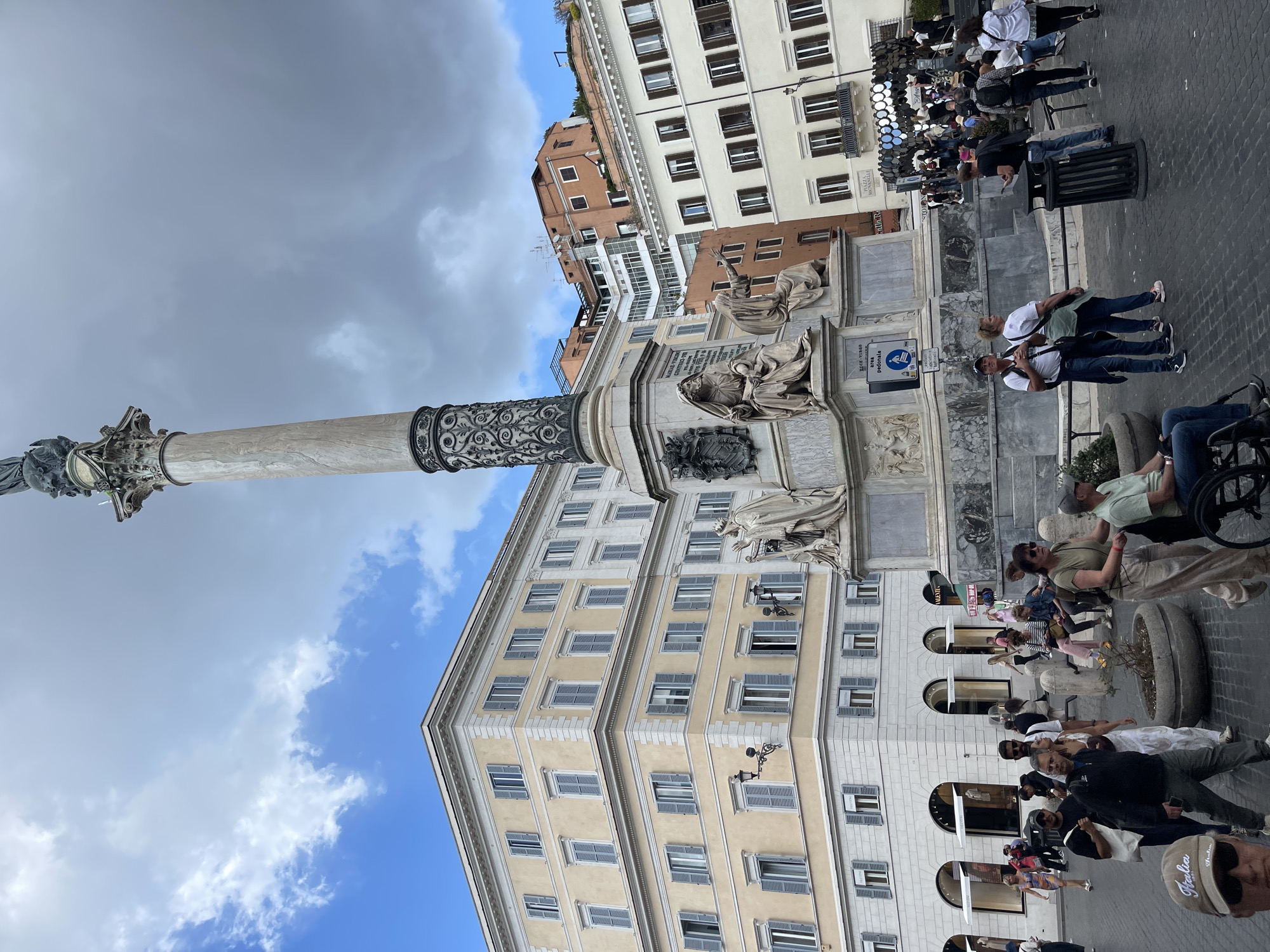 The image depicts the Column of Marcus Aurelius, a Roman monument in Piazza Colonna, Rome, Italy. The column, made of marble, rises prominently against a backdrop of buildings and a partly cloudy sky. The base of the column is adorned with statues and reliefs. The surrounding area is populated with people, some walking, others standing and observing. The buildings around the piazza exhibit classical Roman architecture with multiple stories and shuttered windows. A streetscape stretches along the base of the buildings, lined with shops and pedestrian traffic.