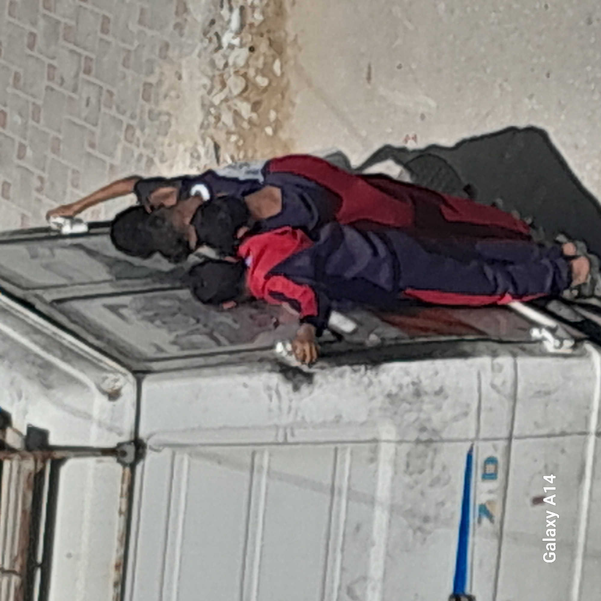 Three individuals are observed on the roof of a white truck. All individuals are wearing clothing comprised of red and blue colors. One individual appears to be holding a white object. The background shows a wall. The location appears to be in an urban environment of the city of Unknown, Unknown. A watermark indicating "Galaxy A14" is visible in the bottom right.