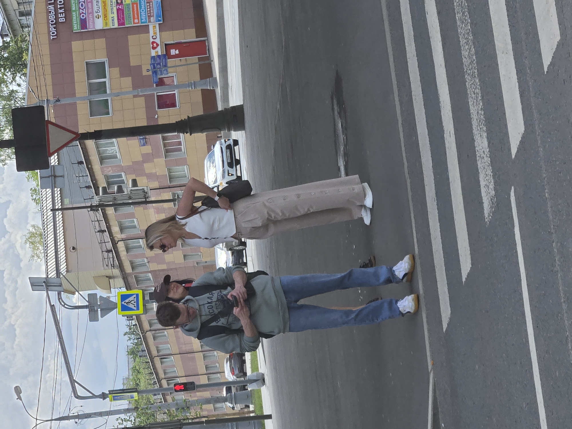 This is an outdoor, medium-range shot taken on a cloudy day. Three people are visible in the foreground, standing on a pedestrian crossing on a city street. A young man in a green hoodie and blue jeans is looking at his phone. A woman in a white vest and beige pants is standing next to him, looking to her right. Another woman, partially visible between them, is wearing a brown baseball cap and appears to be looking at the man. Cars are visible in the background, as are buildings and streetlights. One of the buildings has signs with Russian text. The scene appears to be in Moscow, Russia.
