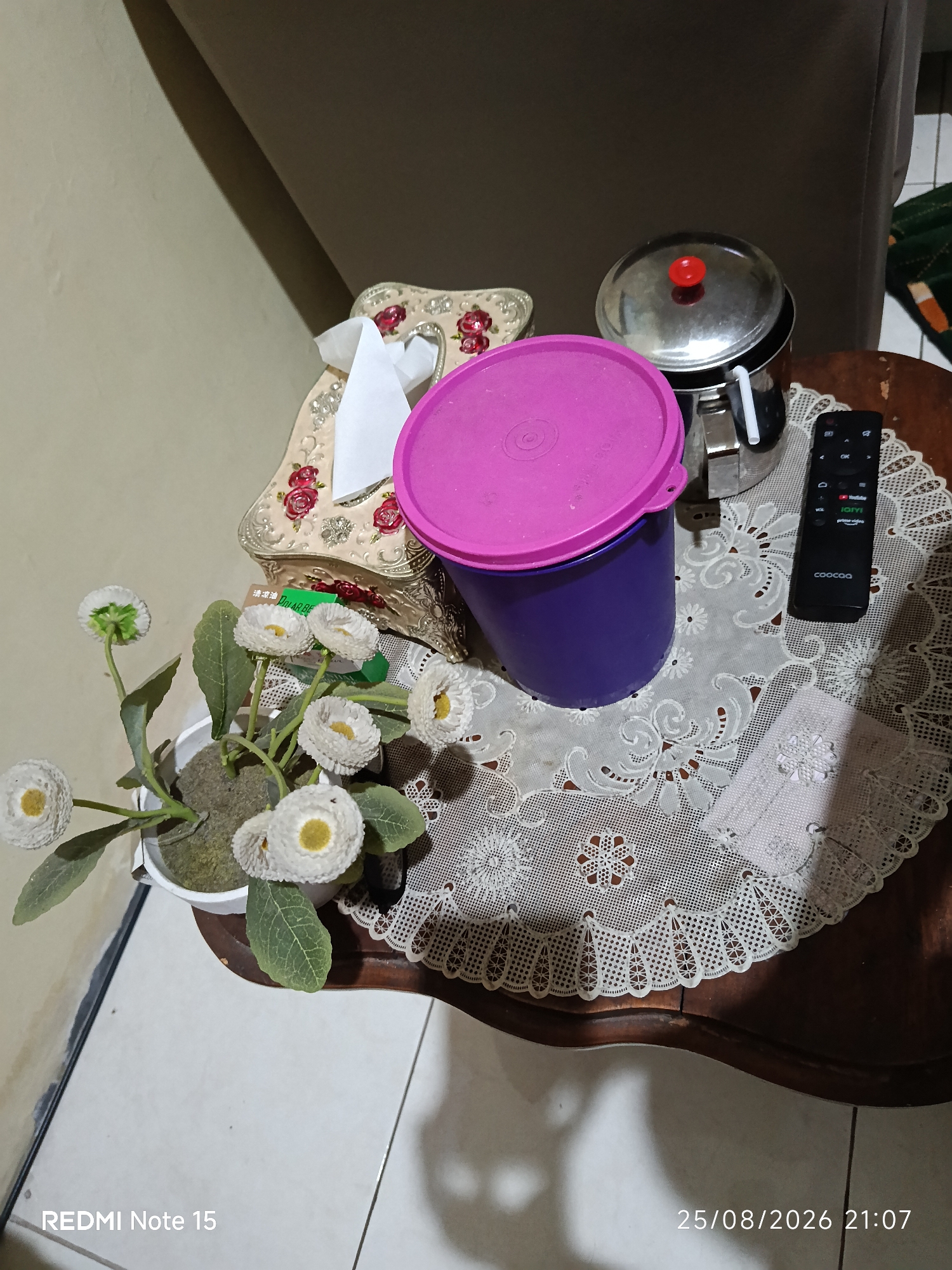 The image depicts a still life arrangement on a wooden table, likely in a domestic setting. The scene is illuminated by artificial light, casting shadows.
In the foreground, a potted artificial plant with several white daisy-like flowers and green leaves is positioned on the left side. The plant is in a white pot filled with a gray granular substance.
To the right of the plant, a tissue box with an ornate, gold-toned frame and red floral embellishments is visible. A white tissue is pulled out from the box.
Next to the tissue box, a tall, purple container with a bright pink lid is present. A straw protrudes from the lid.
Behind the purple container, a stainless steel pot with a red knob on its lid is situated. A straw also appears to be sticking out from this pot.
A black remote control with various buttons is placed on the table to the right of the purple container. The remote has labels such as "VOL," "OK," and icons for streaming services like YouTube and Prime Video. The brand name "coocaa" is visible at the bottom of the remote.
A white, lacy tablecloth covers a portion of the wooden table.
In the background, a portion of a beige wall and a dark-colored sofa are visible.
The timestamp "25/08/2026 21:07" is present in the bottom right corner of the image, along with the text "REDMI Note 15" in the bottom left corner.
The provided image does not contain enough information to determine its specific location within the city of Keboananom, Indonesia. However, the presence of household objects suggests it is likely taken in a residential area.