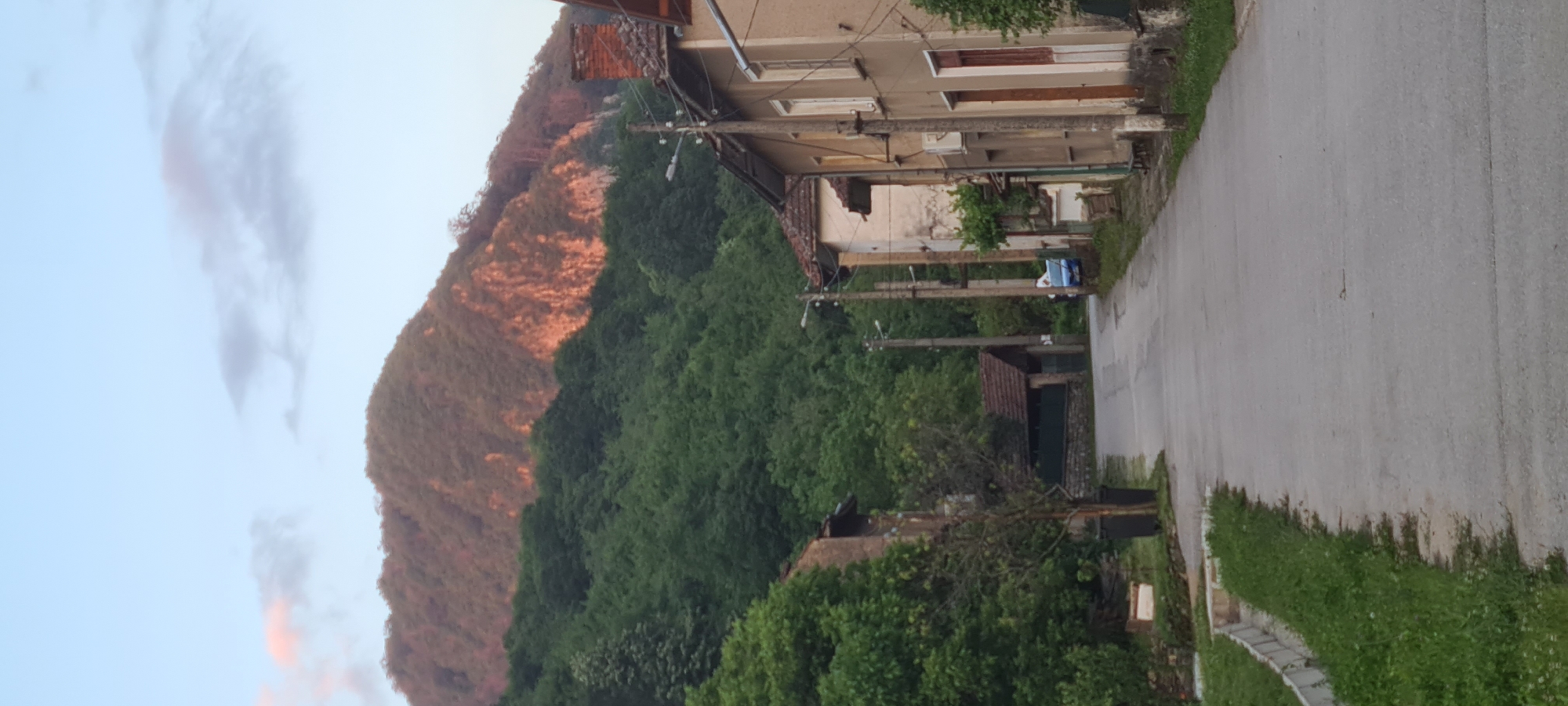 The image depicts a rural village street in Lopian, Bulgaria, at either sunrise or sunset, with a prominent mountain in the background. The sky is light blue with scattered, pale clouds, some exhibiting pink and orange hues.
In the background, a large mountain dominates the upper half of the image. Its upper slopes feature areas of exposed rock and sparser, reddish-brown vegetation, strongly illuminated by a warm, orange-red light. The lower slopes of the mountain are densely covered with vibrant green deciduous trees.
A paved asphalt road forms the central element of the foreground and midground, curving gently towards the left as it recedes into the distance towards the mountain. The road surface appears somewhat worn and possibly damp.
On the left side of the road, a narrow grass verge with concrete curbing runs parallel to the pavement. Further back, a small, bare tree stands next to two dark waste bins. Behind these, a two-story building with a light brown facade and a dark roof is partially visible, mostly obscured by trees. Further along the road on the left, a smaller, rustic building with a terracotta tiled roof and a stone foundation is seen.
On the right side of the road, a series of traditional two-story residential buildings line the street. These structures feature varying shades of beige and light brown facades, some showing signs of age and wear, and are topped with terracotta tiled roofs. Multiple wooden utility poles with overhead wires are positioned along this side, extending down the street. A small blue car is partially visible, parked on the right side of the road behind one of the utility poles. An outdoor air conditioning unit is mounted on the side of one of the buildings, and a brick chimney is visible on the roof of the closest building on the right.
No people or animals are visible in the scene. The overall impression is one of a quiet, inhabited rural landscape.