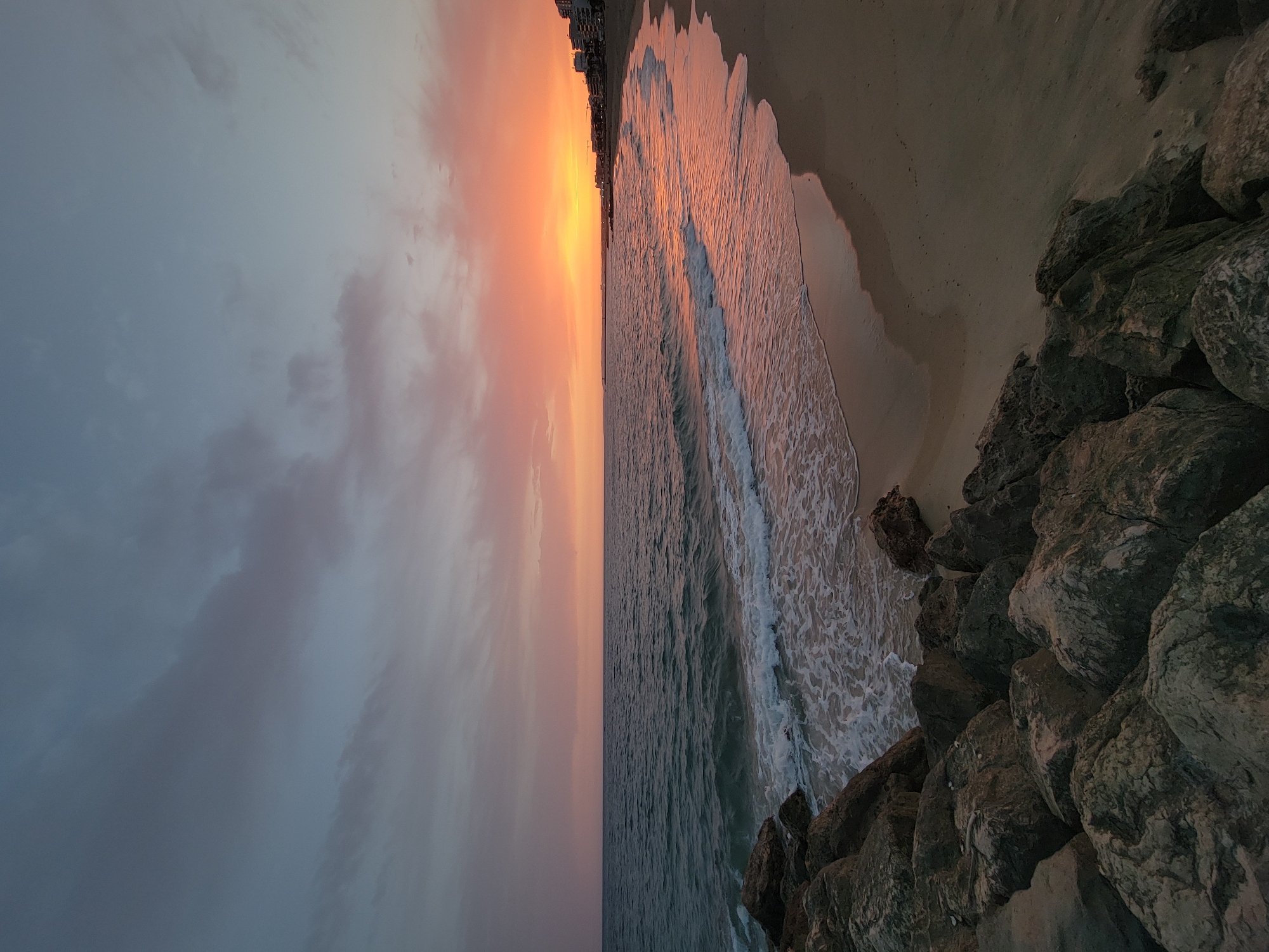 This is a landscape photograph taken at a beach in Quarteira, Portugal, during sunset. The image is oriented vertically, showing a view from a rocky outcrop looking out towards the ocean and the sky.
The subjects are the natural elements of the scene. The foreground is dominated by a pile of large, dark grey rocks. Adjacent to the rocks is a sandy beach, wet from the receding tide, with gentle waves breaking on the shore. The ocean occupies a significant portion of the frame, its surface textured with ripples and frothy whitecaps. The sky above is a dramatic canvas of color, transitioning from a soft, muted grey with hints of pink and purple on the left, to a vibrant gradient of orange and yellow near the horizon on the right, where the sun is setting. The warm hues of the sunset reflect on the wet sand and the surface of the ocean. In the far distance, along the horizon, a line of buildings suggests a coastal town.
The time of day is clearly sunset, with the characteristic warm light and changing colors in the sky. The weather appears calm, with gentle waves indicating tranquil conditions. The presence of the ocean, sand, rocks, and buildings confirms the coastal setting of Quarteira, Portugal. The overall mood conveyed by the image is one of peacefulness and natural beauty, enhanced by the serene ambiance of the sunset. There is no visible text in the image.