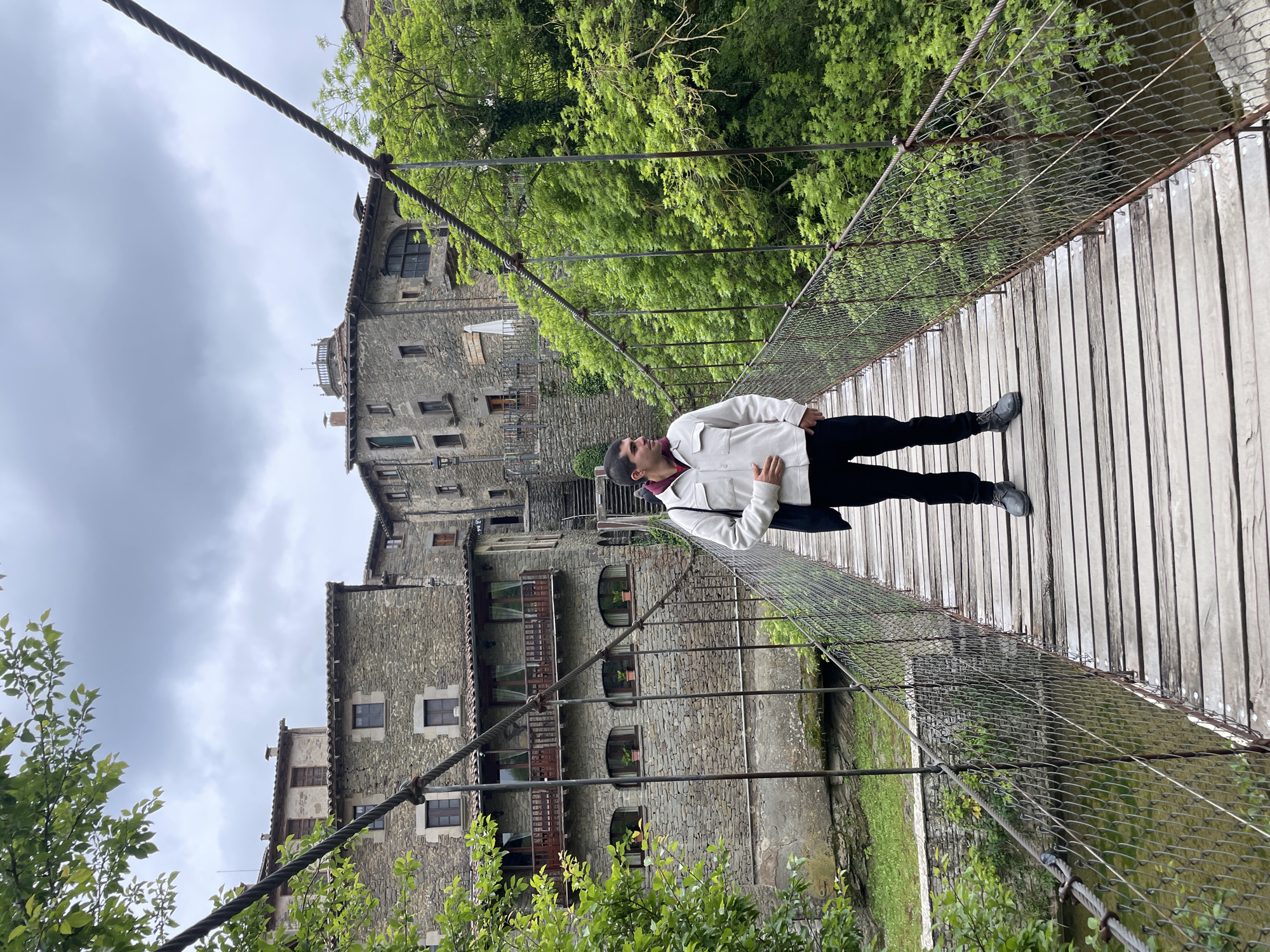 The image shows a person standing on a wooden suspension bridge in Rupit i Pruit, Spain. The individual is wearing a white jacket, black pants, and dark shoes. They are looking upwards and have a bag slung over their shoulder. The bridge has metal cables and netting on the sides. Behind the person, there are old stone buildings and green trees. The sky appears cloudy. The perspective suggests the photo was taken from a low angle.