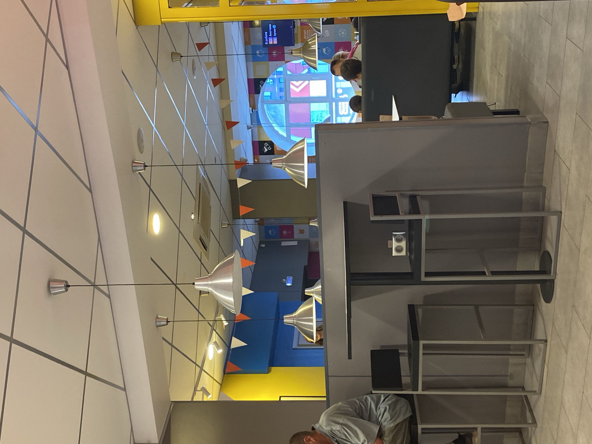 The image shows an interior space with a modern design. The walls are light with grey linear patterns. Several pendant lights with conical shades hang from the ceiling. Bunting with triangular flags in white, orange, and red is strung along the walls. In the background, there is a circular window with colorful panels. Several people are present, though their faces are not clearly visible. A man is visible in the foreground, and others are standing near the window. The room contains shelves and furniture, suggesting a public space. The overall setting appears to be in a modern, possibly educational or cultural center.
The presence of Cyrillic text in the background and the modern design style may suggest the location is Votkinsk, Russia, but without explicit confirmation, this is inferential.