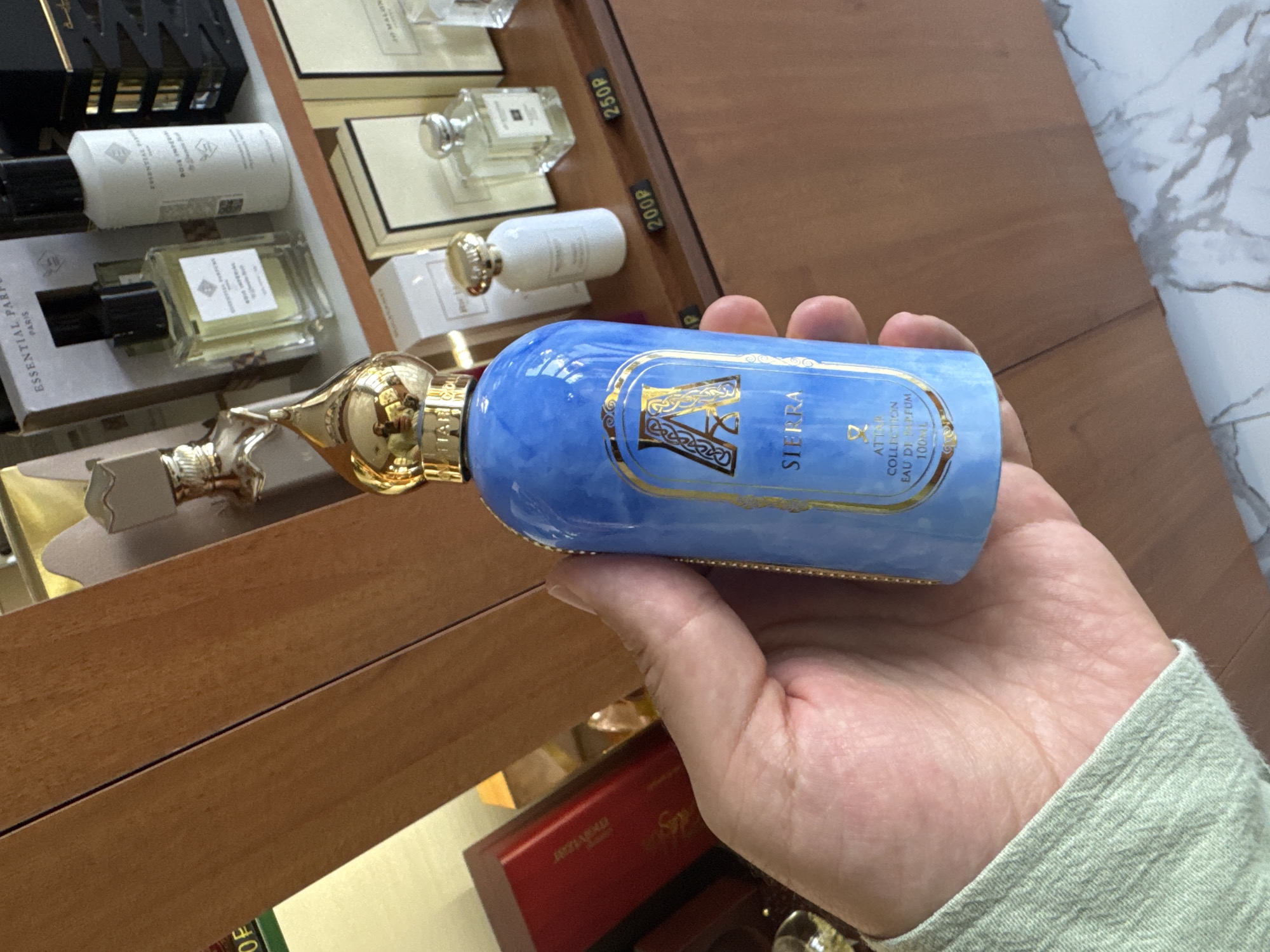 A hand holds a blue perfume bottle with a gold cap and a gold label that reads "SIERRA COLLECTION EAU DE PARFUM 100ML" in Krasnodar, Russia. The background shows shelves filled with various perfume bottles and boxes. The lighting is bright, and the overall scene appears to be inside a retail store.