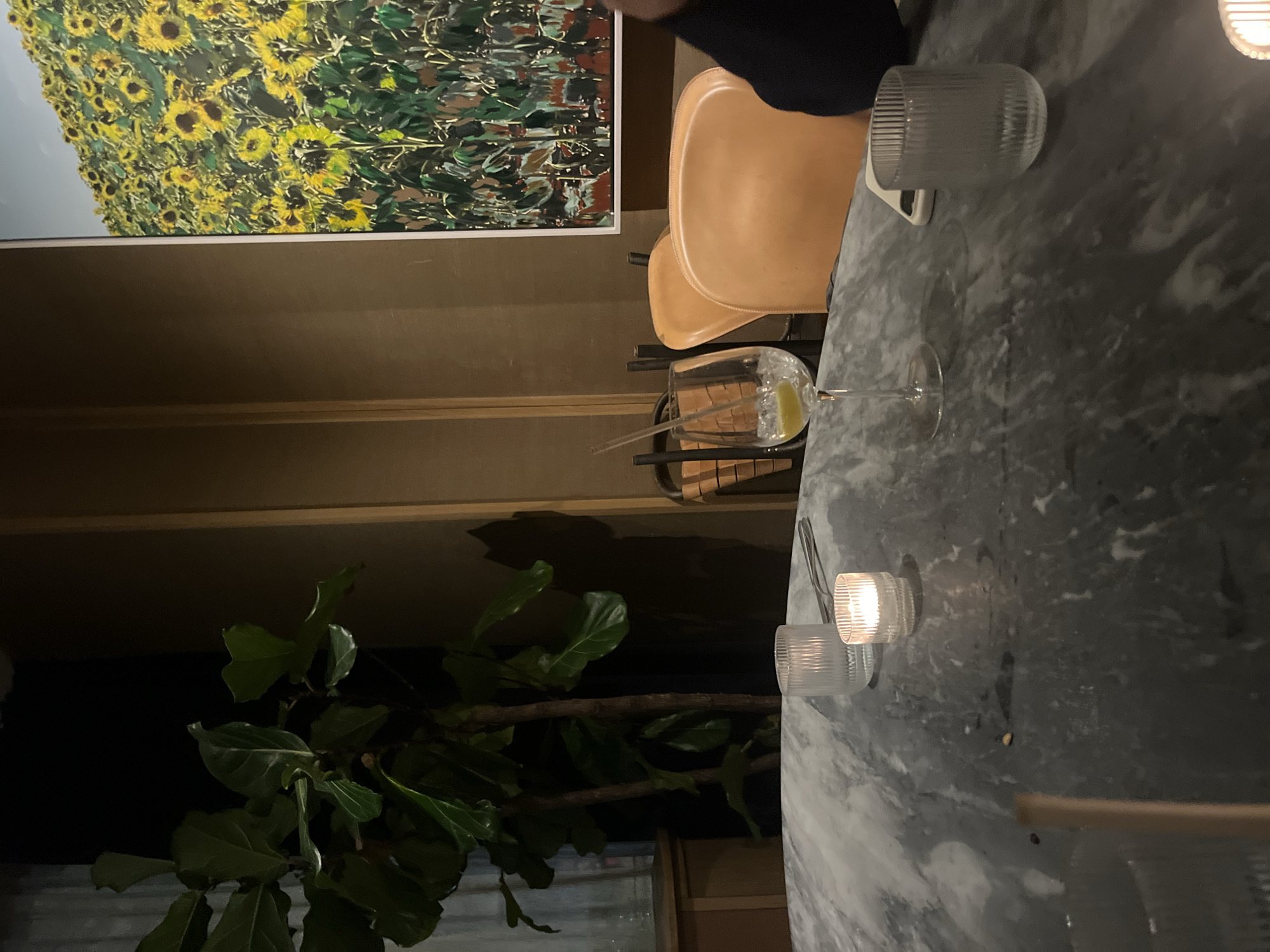 The image portrays an intimate indoor setting, likely a restaurant or bar in Paris, France, during the evening or night. A dark grey marble table with white veining occupies the right side of the frame. On its surface, a stemmed glass holds a clear beverage with a lime slice and two straws. Another clear, ribbed glass is placed further up the table, and two small, ribbed glass candle holders emit a warm, soft light from the bottom right. A person's arm, dressed in dark clothing, is partially visible at the top edge of the table.
The left side of the scene features a wall adorned with a large, framed painting depicting a lush field of yellow sunflowers and green foliage against a lighter background. Below the artwork, a light brown wall paneling sets the background for two modern, light brown chairs with distinctive curved backs. In the lower-left corner, a large green plant with broad leaves, possibly a Fiddle-leaf Fig, thrives in the ambient light. The scene evokes a cozy, sophisticated, and relaxed atmosphere. No visible text is present.