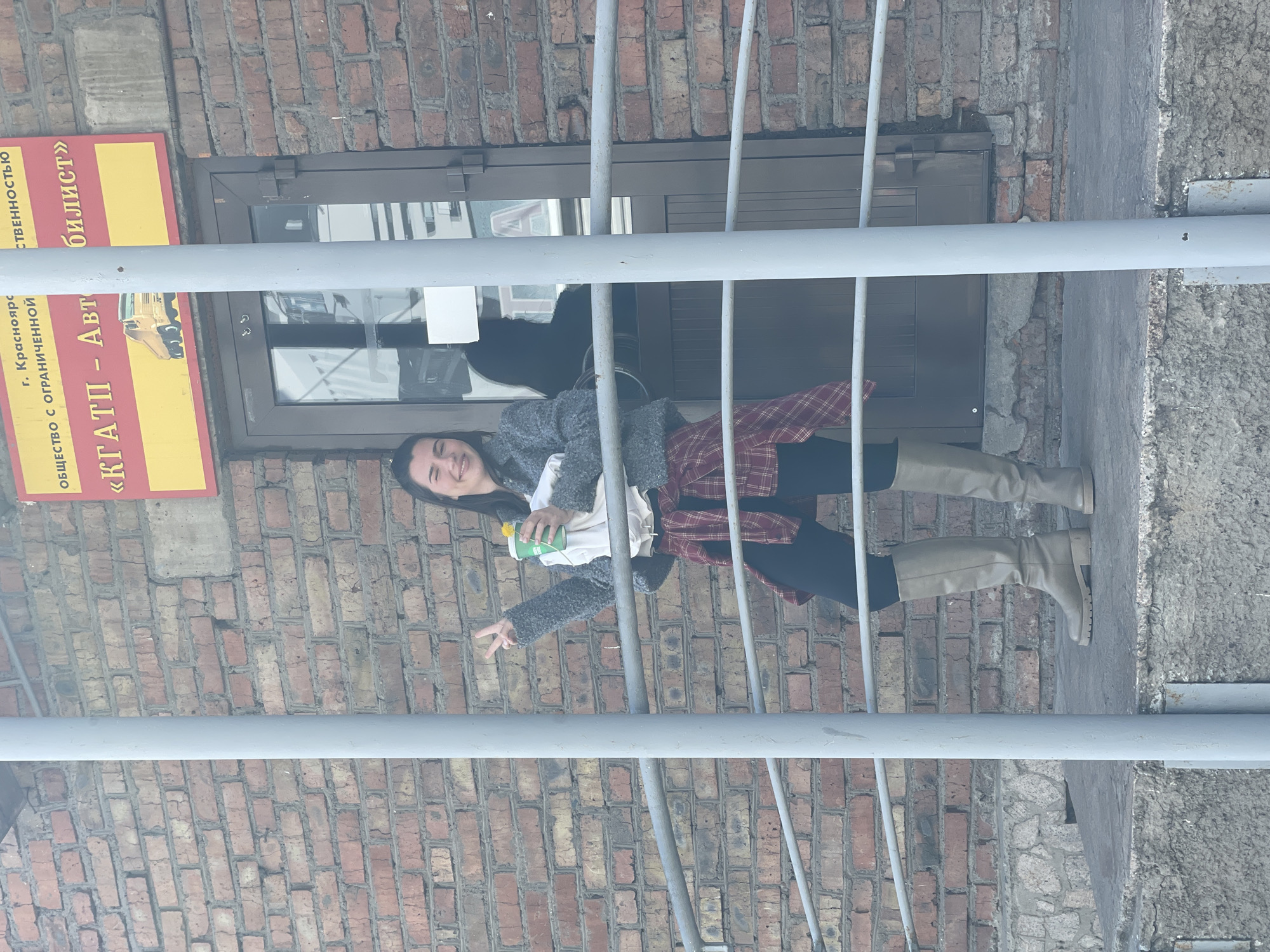 A woman is standing on stairs in front of a brick building. She is wearing a gray sweater, a red plaid shirt tied around her waist, dark pants, and tan boots. She holds a green bottle in one hand and makes a peace sign with the other. A sign reading "Krasnoyarsk Autotransport Enterprise" in Russian is affixed to the building. The location is Krasnoyarsk, Russia. The stairs are metal and weathered. The overall scene appears to be outside.