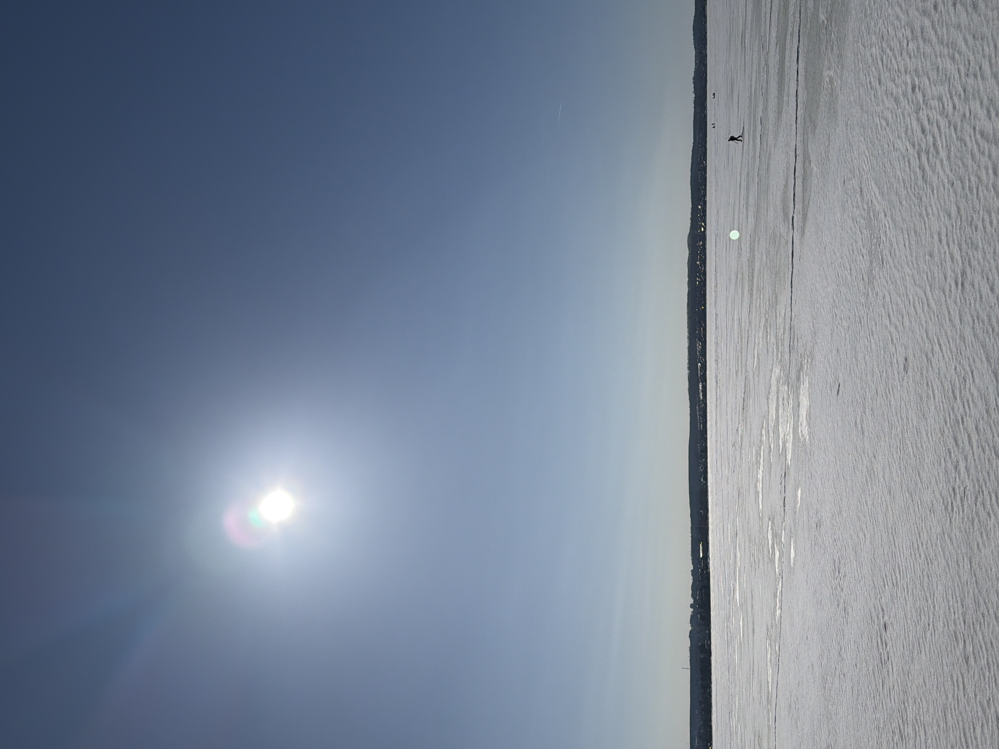 The image displays a panoramic view of the frozen Votkinsk Pond in Votkinsk, Russia, under a clear, bright blue sky. The upper two-thirds of the frame are dominated by the sky, featuring a prominent, bright sun in the upper-left quadrant, which produces noticeable lens flare with green and purple chromatic aberrations. A faint white contrail is visible in the upper-right sky.
A dark, low-lying horizon line stretches across the mid-ground, representing the distant shoreline of Votkinsk. This silhouette reveals a series of structures and trees, with a distinct tall, thin vertical structure visible on the left side of the cityscape.
The lower third of the image consists of the vast, snow-covered surface of the frozen pond. The surface exhibits variations in texture, with areas of rippled snow and smoother, more reflective ice patches. A distinct horizontal line or crack in the ice extends across the mid-ground. A single human figure, visible as a dark silhouette, walks or skates on the frozen surface in the mid-right portion of the frame, moving away from the viewer. Several smaller, less defined dark specks, potentially other individuals or objects, are visible further in the distance on the ice towards the right.