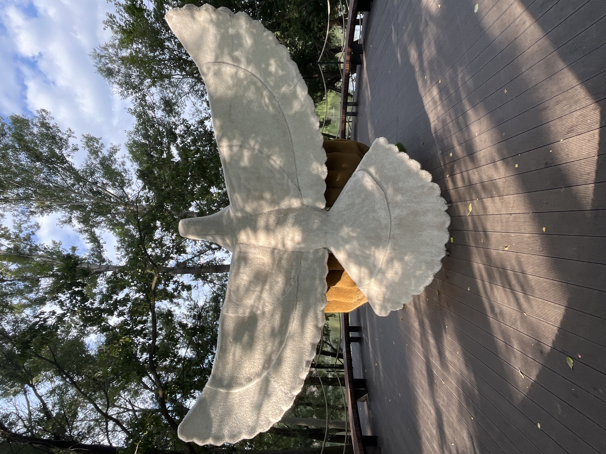 The image shows a large, white, stylized sculpture of a bird with its wings outstretched. The sculpture is positioned on a wooden deck in an outdoor setting. Behind the sculpture, there are trees and a body of water, possibly a river, with a rope railing and benches along the edge. The sky is visible above, with clouds and sunlight filtering through the trees, casting shadows on the deck. The sculpture appears to be made of a soft, textured material. This artwork is located in Blagoveshchensk, Russia, specifically in the Amur Bay area, near the embankment. There are no people or animals visible in the frame.