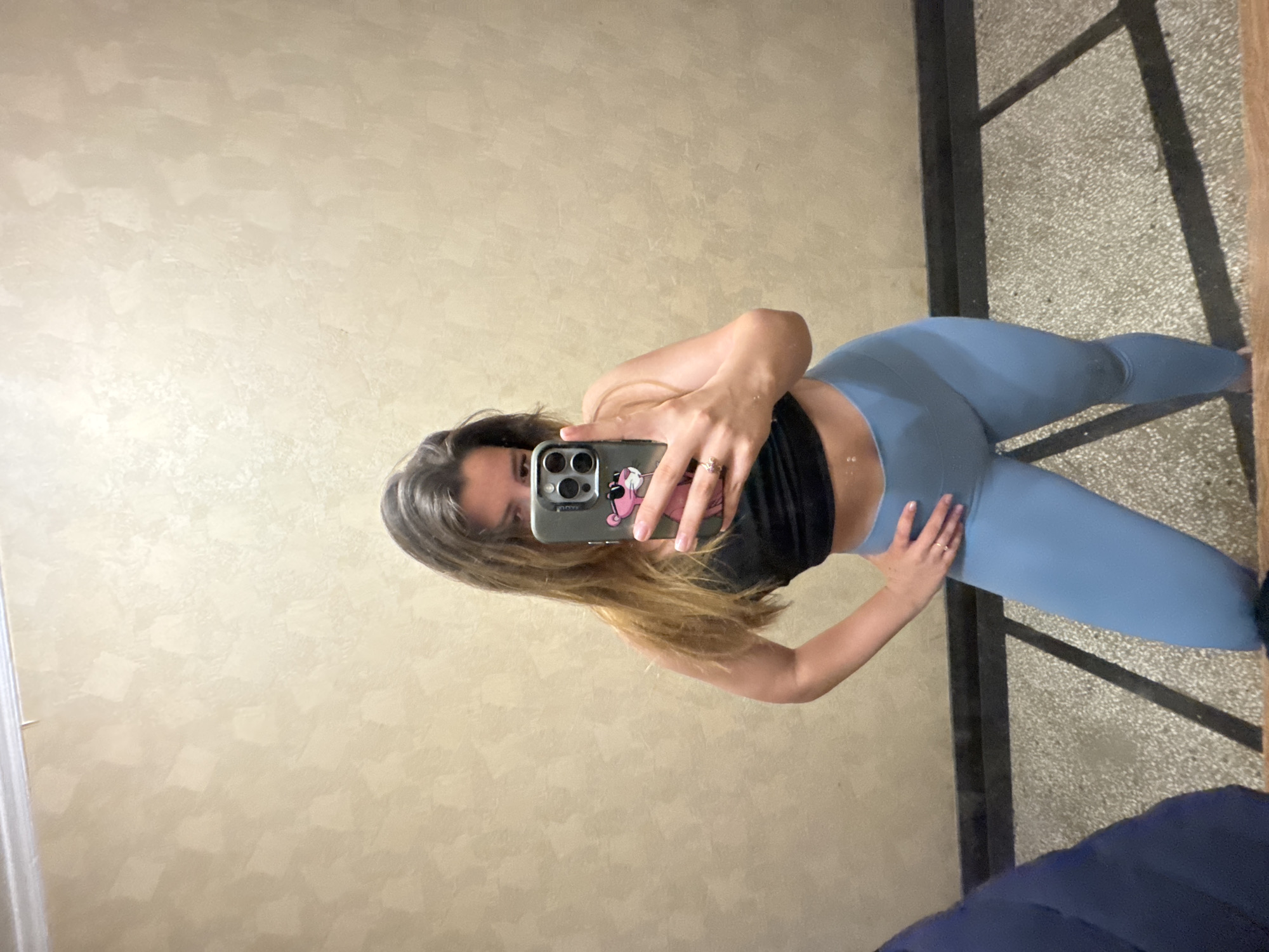 The image displays a mirror selfie of a woman in an indoor setting in Sofia, Bulgaria. She is positioned facing the camera, which is her smartphone, held up to obscure part of her face. She has long, dark brown hair and is wearing a black crop top or sports bra paired with light blue, form-fitting leggings. On her right hand, a ring with a light-colored stone is visible. Her smartphone case features a pink cartoon character, possibly Minnie Mouse, and the partially visible text "AQU".
The background reflects a light beige wall with a textured, possibly stucco or Venetian plaster finish. To the right, a section of a dark-colored structure, resembling a railing or partition, is visible, extending down to a light-colored, speckled floor. Another dark blue item, likely a piece of clothing, can be seen in the lower right corner, resting on the floor. The lighting appears to be artificial, typical of an indoor environment. The woman's visible eye suggests a casual or neutral expression while posing for the photo.